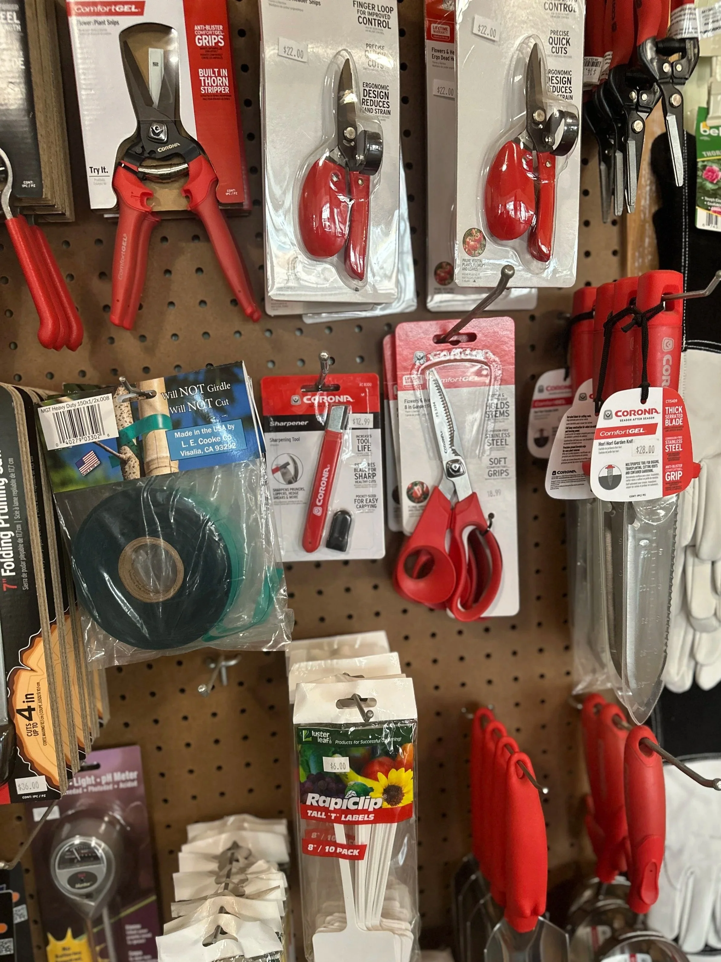 A display of various gardening and household tools including pruning shears, scissors, a tape measure, plant labels, and gloves, hanging on a pegboard in a store.