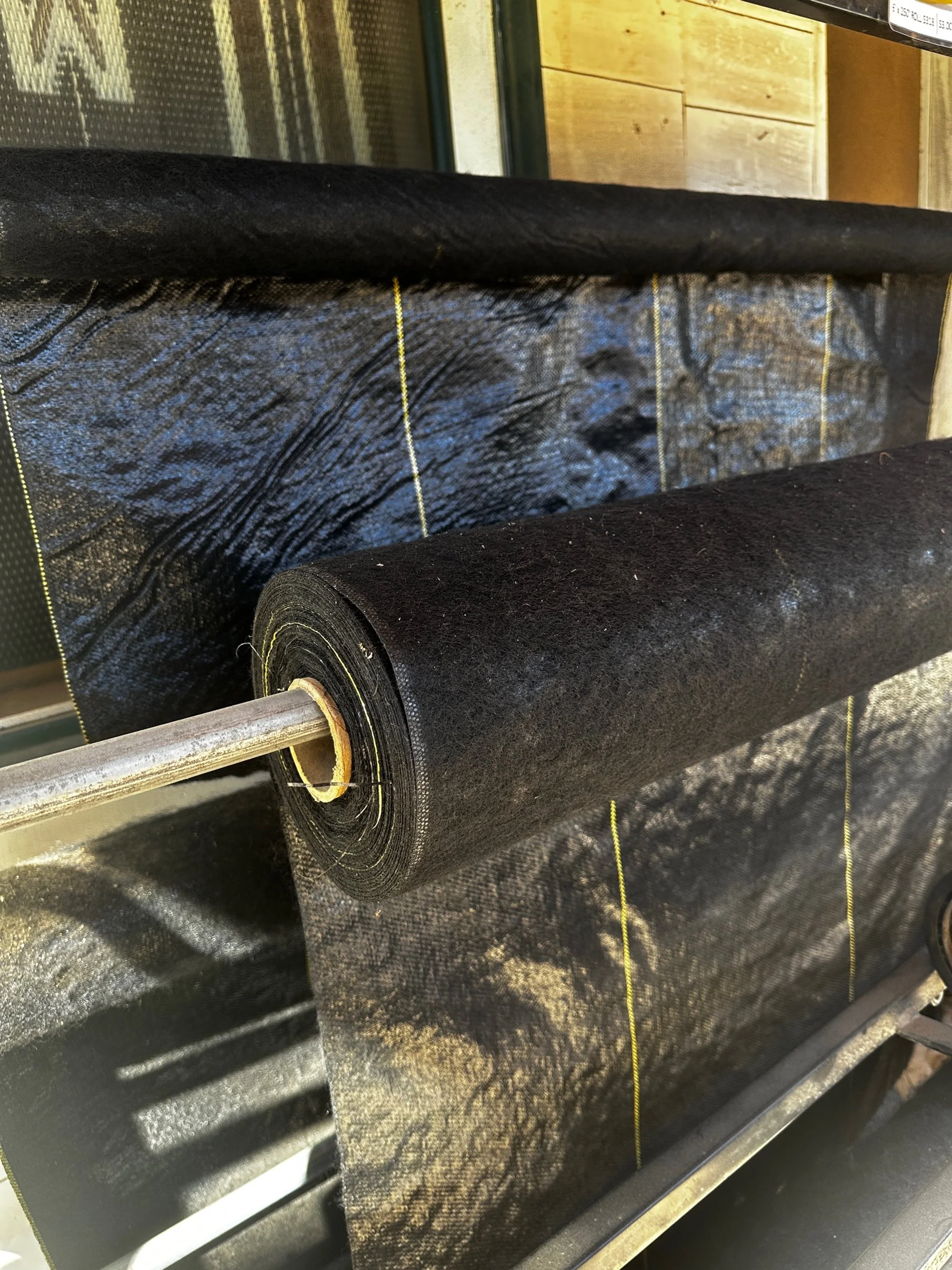 Roll of black material, possibly roofing or waterproofing membrane, on a metal tube in an industrial or construction setting.