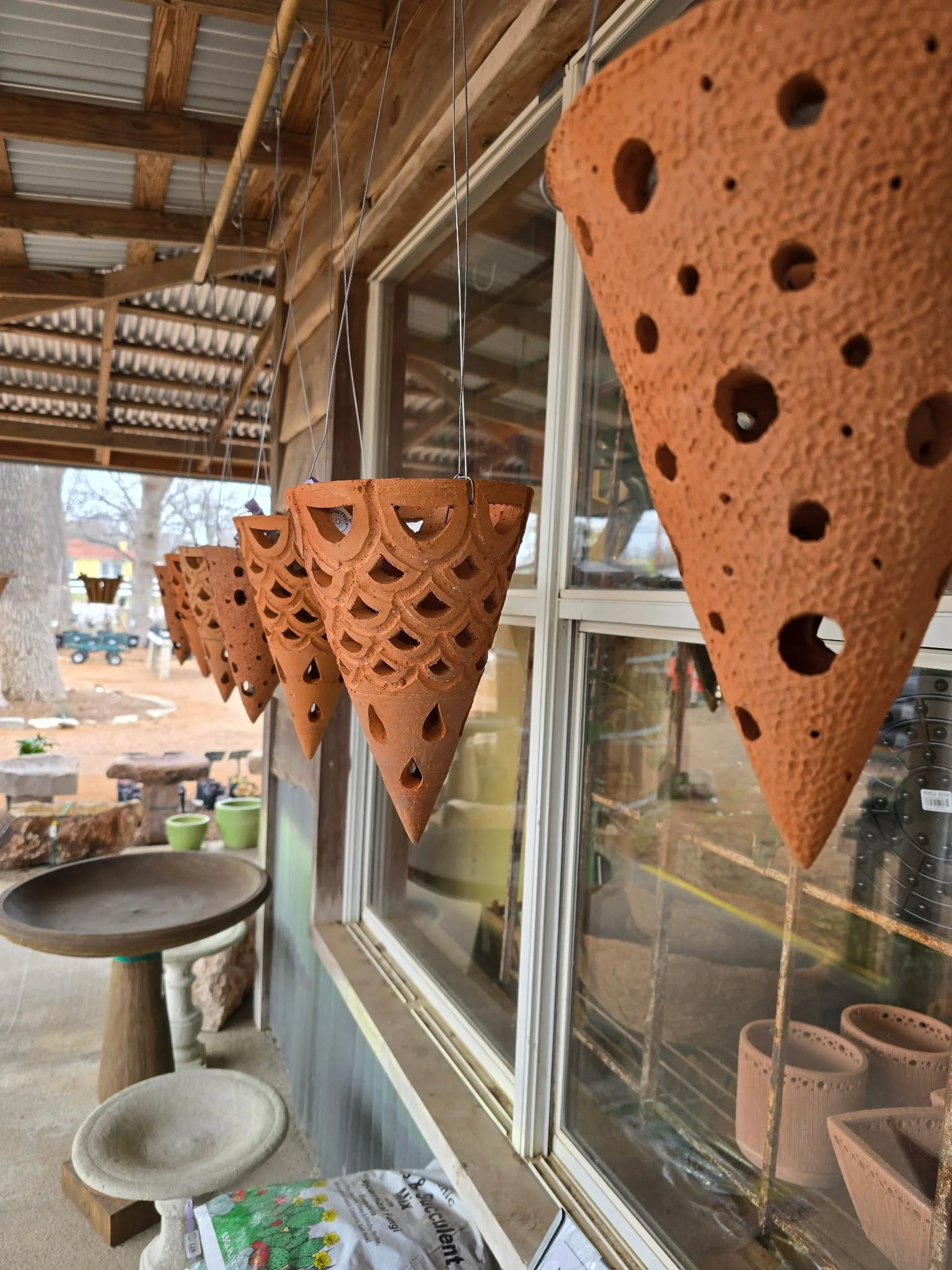 Terracotta hanging planters with decorative cutouts, suspended outside a wooden building with large windows, on a porch.
