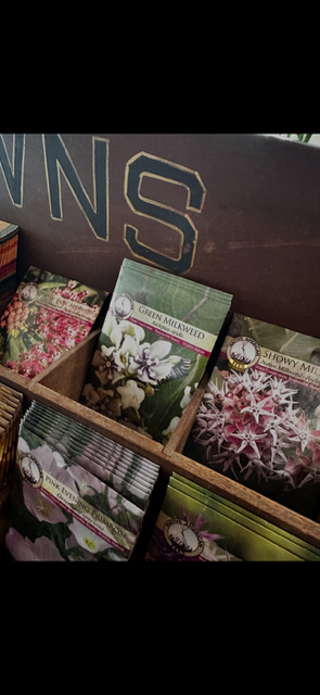 Seed packets for green milkweed, showy milkweed, and other flowering plants inside a wooden display box.