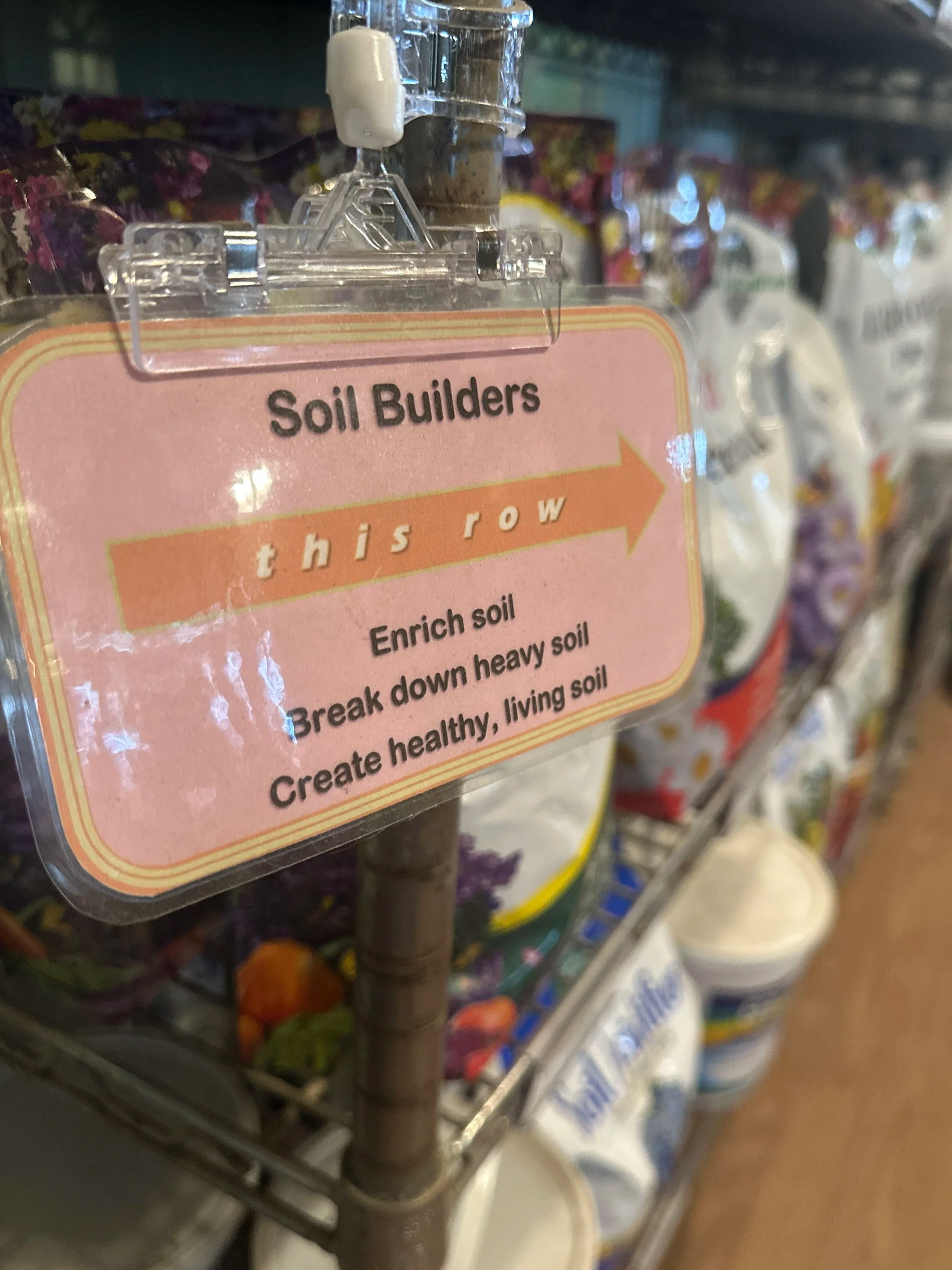 Close-up of a pink sign with an orange arrow reading "this row," instructing to enrich soil, break down heavy soil, and create healthy, living soil, in a store aisle with gardening products.