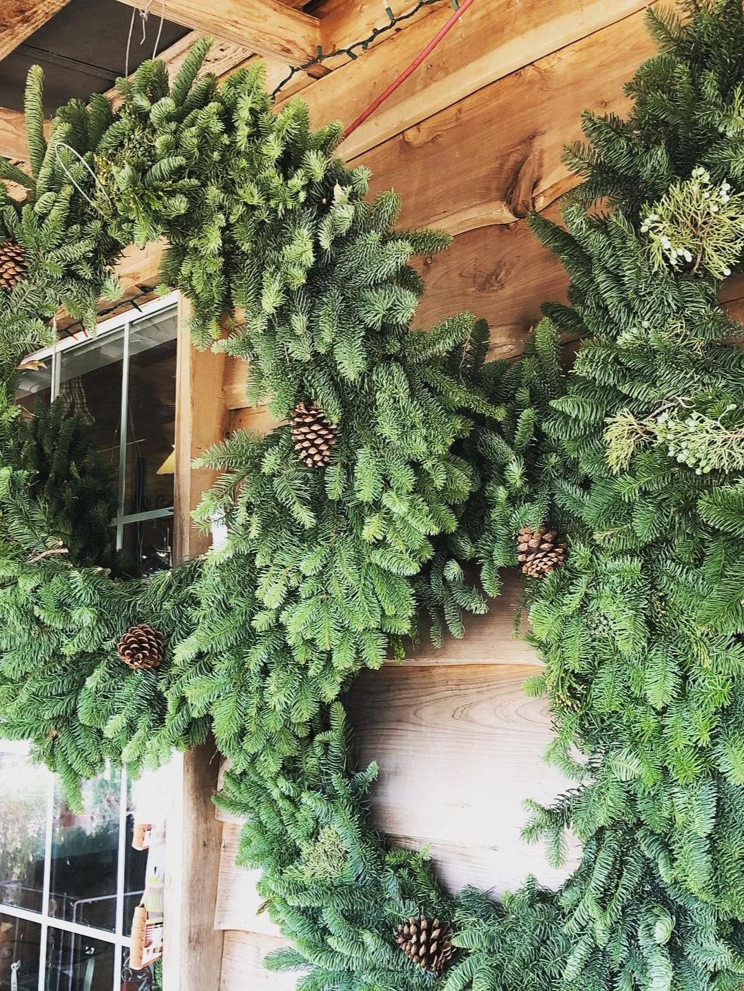 Christmas wreath made of evergreen branches with pinecones hanging on a wooden wall, partially reflecting in a mirror.
