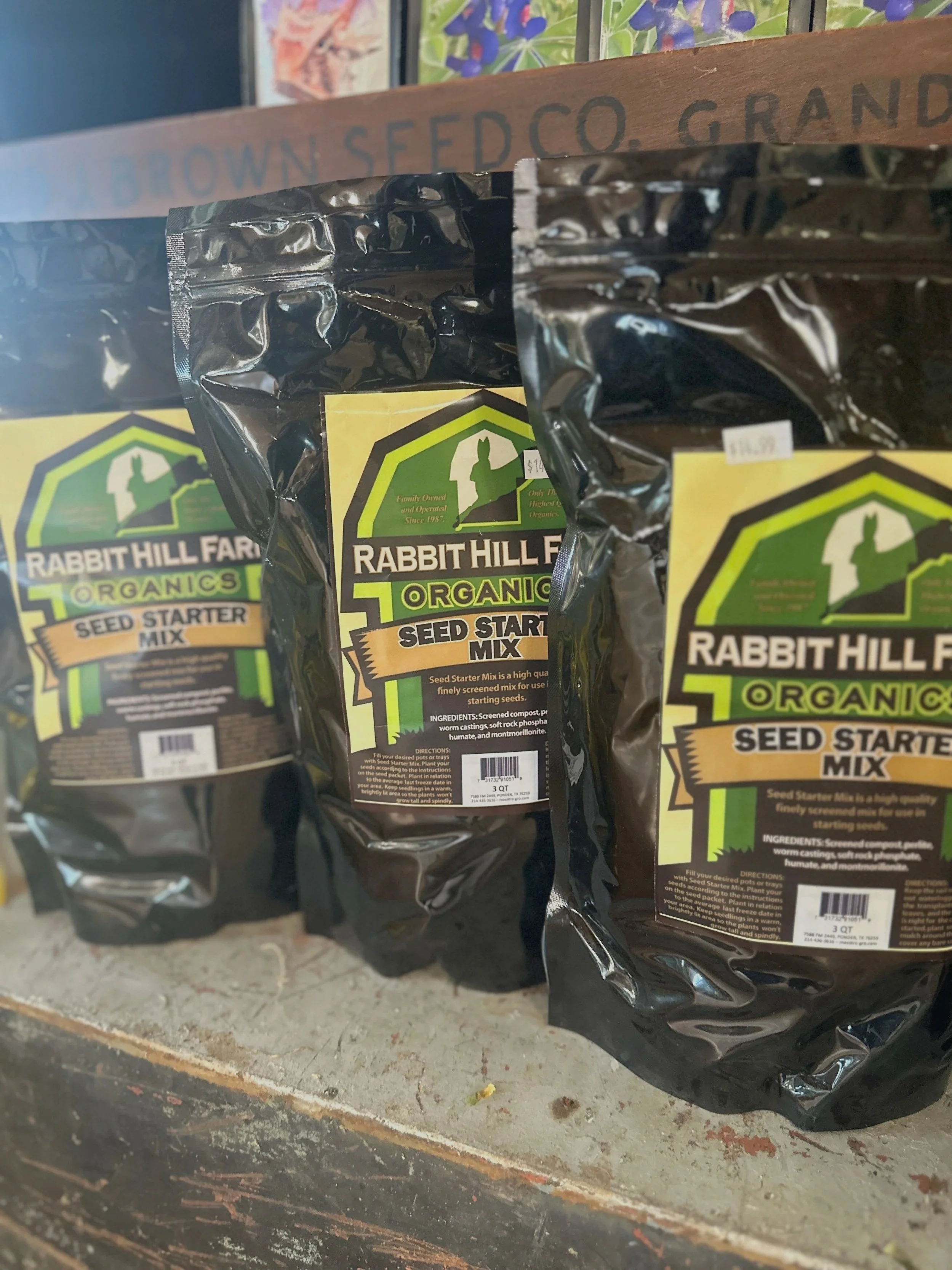 Three black packages of Rabbit Hill Farms organic seed starter mix on a rustic wooden surface, with a colorful artwork or poster in the background.