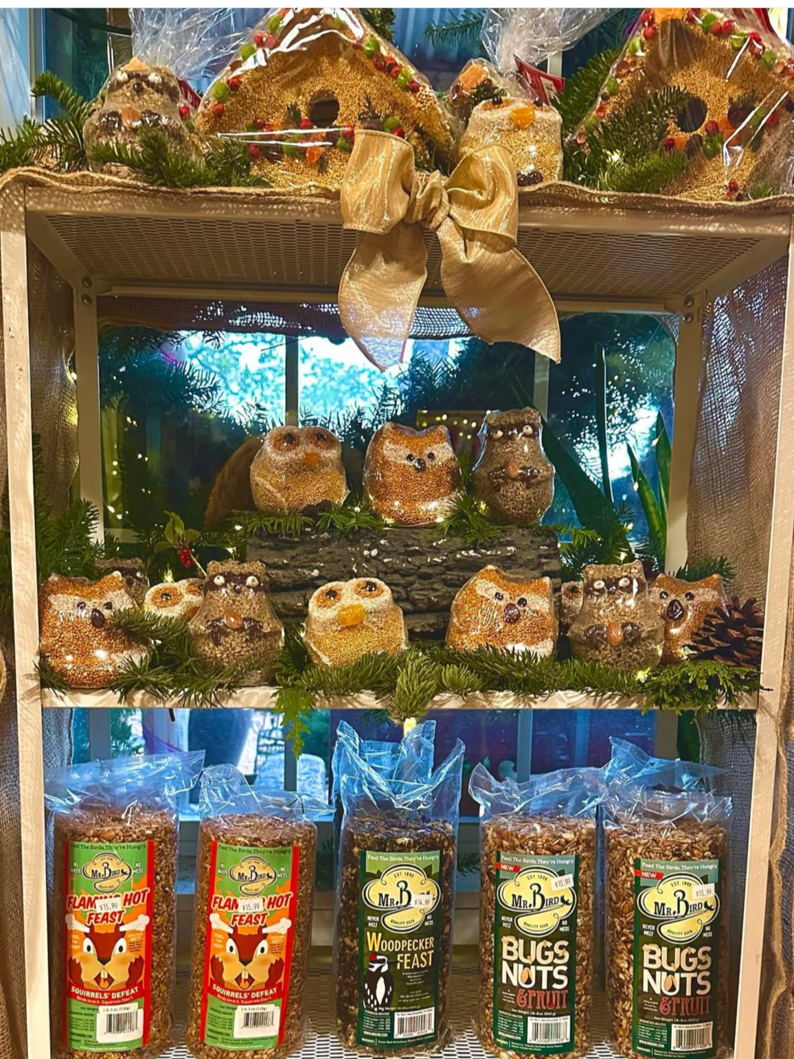 Display of decorative squirrel feeder snacks and seed packages, with squirrel-shaped treats on the shelf, greenery, pinecones, and a large gold ribbon at the top.