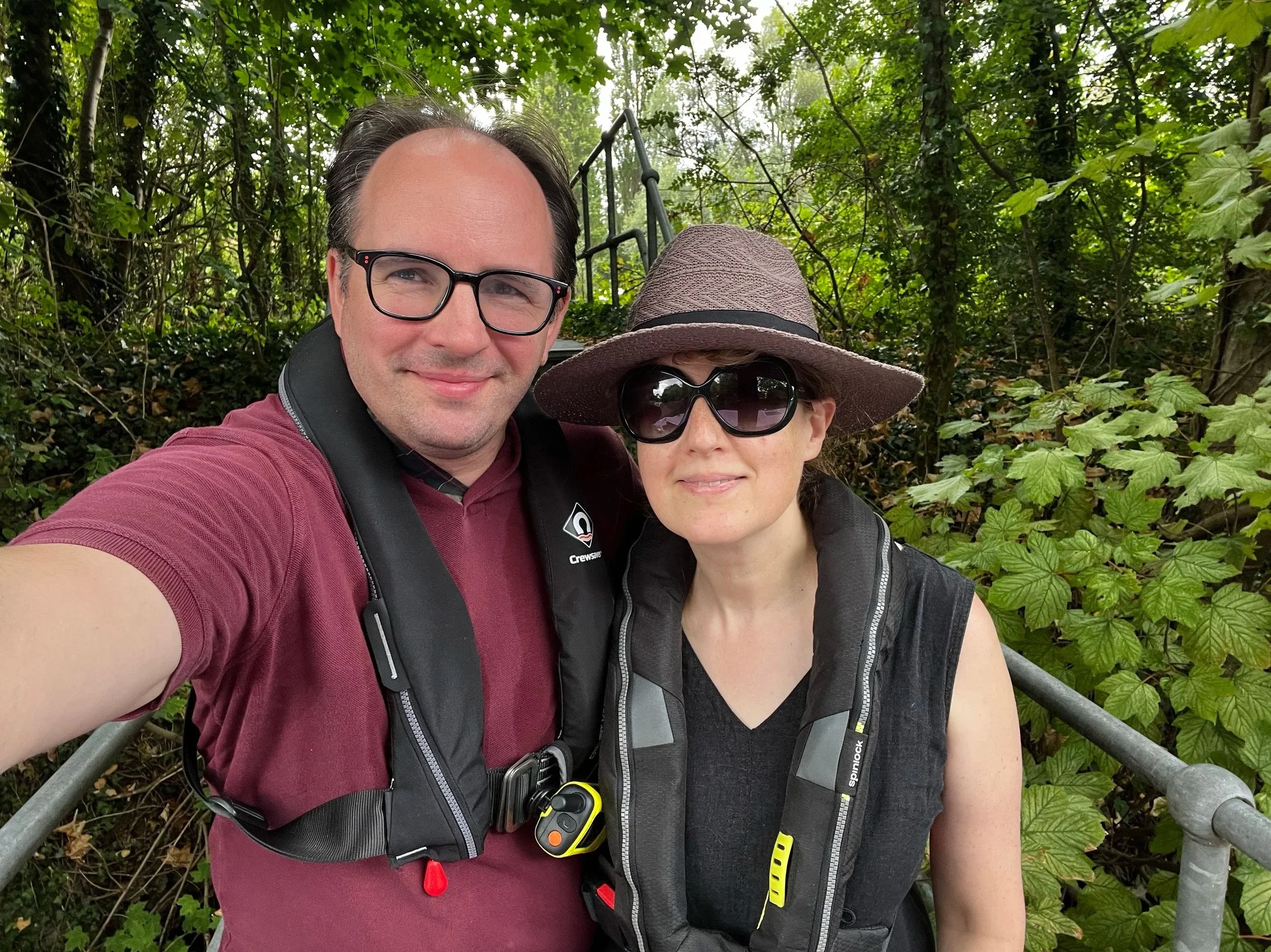 A man and woman taking a selfie outdoors in a green, wooded area. The man is wearing glasses, a maroon shirt, and a black safety harness with a yellow device attached. The woman is wearing large sunglasses, a wide-brimmed hat, and a black vest with safety gear. They are smiling and appear to be enjoying a hike or outdoor adventure.