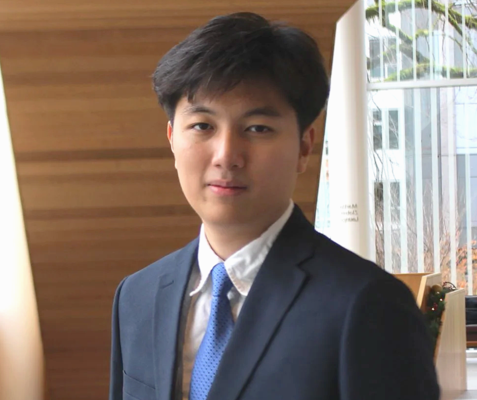 Young man in a dark suit with a blue tie, standing indoors near a wooden wall, with a window behind him.