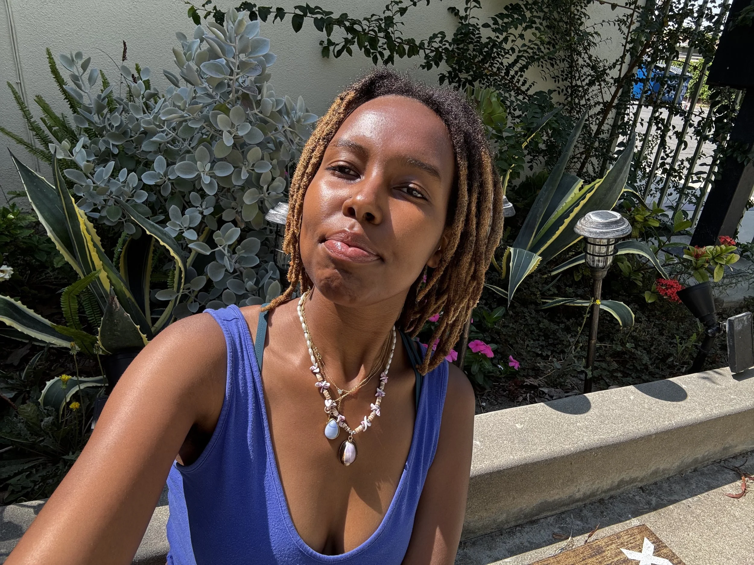 A woman with dreadlocks and tan skin outdoors in sunlight, wearing a blue tank top and layered necklaces, standing in front of various green plants and garden lights.