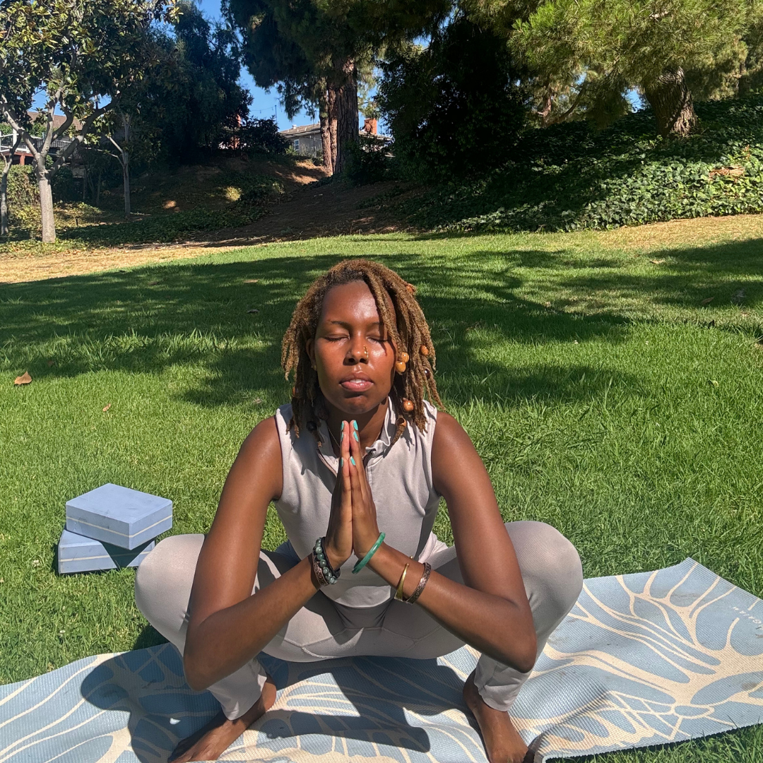 A woman with dreadlocks practicing yoga outdoors on a mat in a grassy park, sitting in a squatting position with hands in prayer pose and eyes closed.