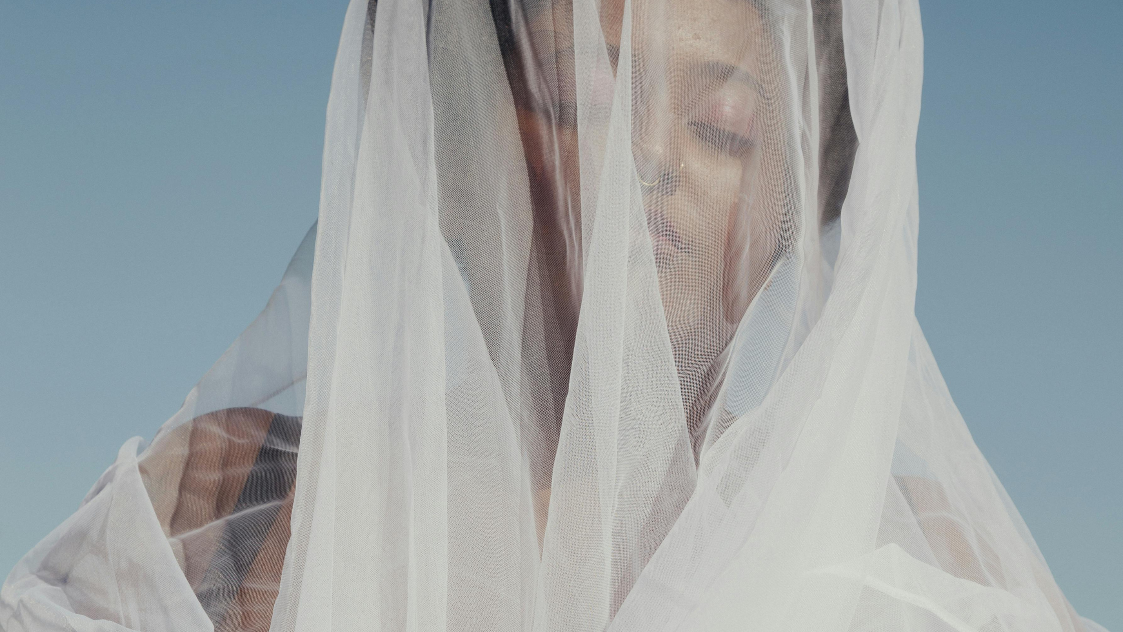 A woman with closed eyes, golden nose ring, and makeup, covered with a sheer white fabric against a blue background.