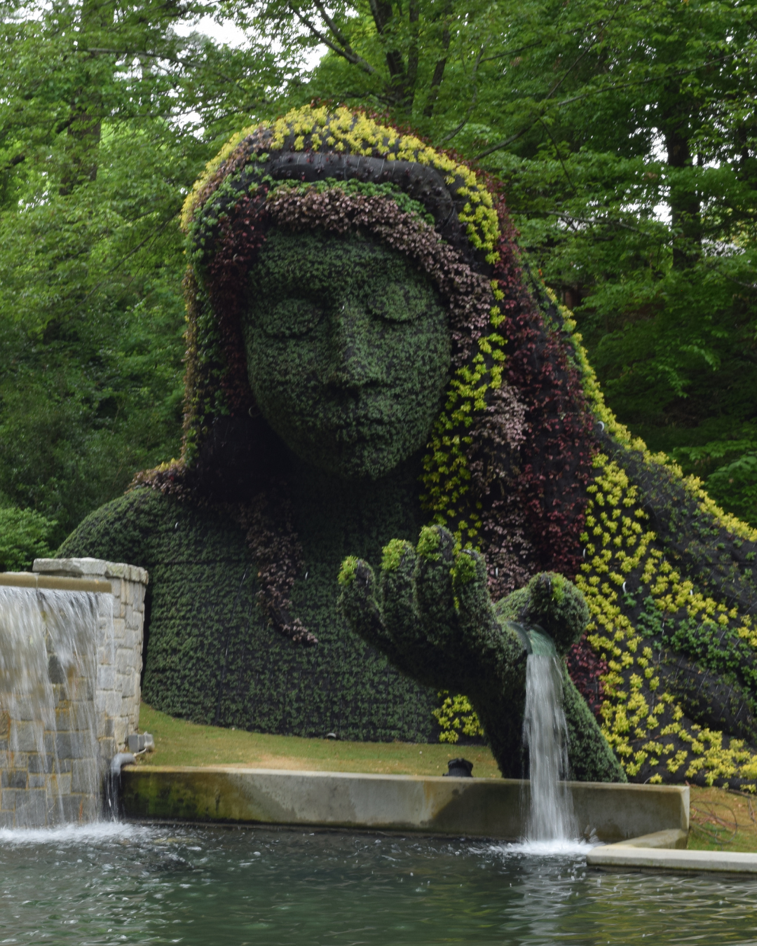 A large garden sculpture of a woman with long hair and closed eyes, covered in green plants and flowers, with water flowing from her hand into a pool.