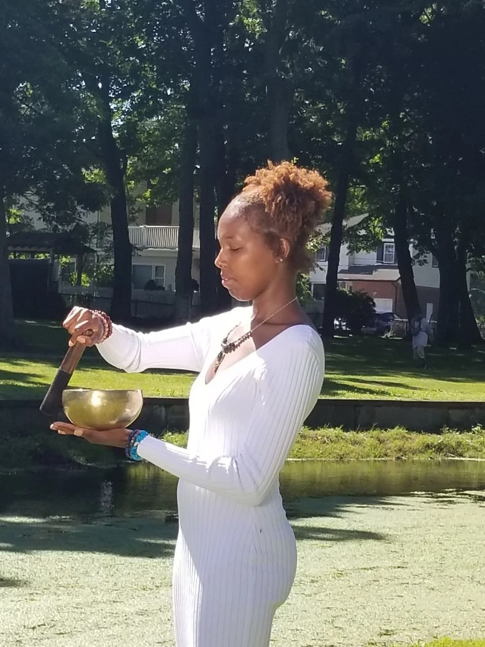 A woman in a white dress holding a singing bowl while using a wooden mallet outdoors in a park with green trees and a pond.