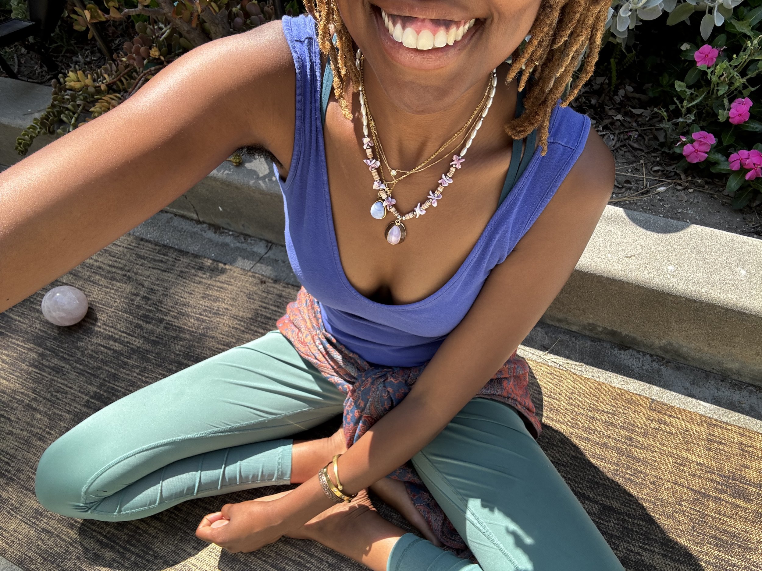 A woman with dreadlocks, wearing a blue sleeveless top, turquoise pants, and layered necklaces, is sitting cross-legged on a wooden bench outdoors, smiling, with pink flowers in the background.