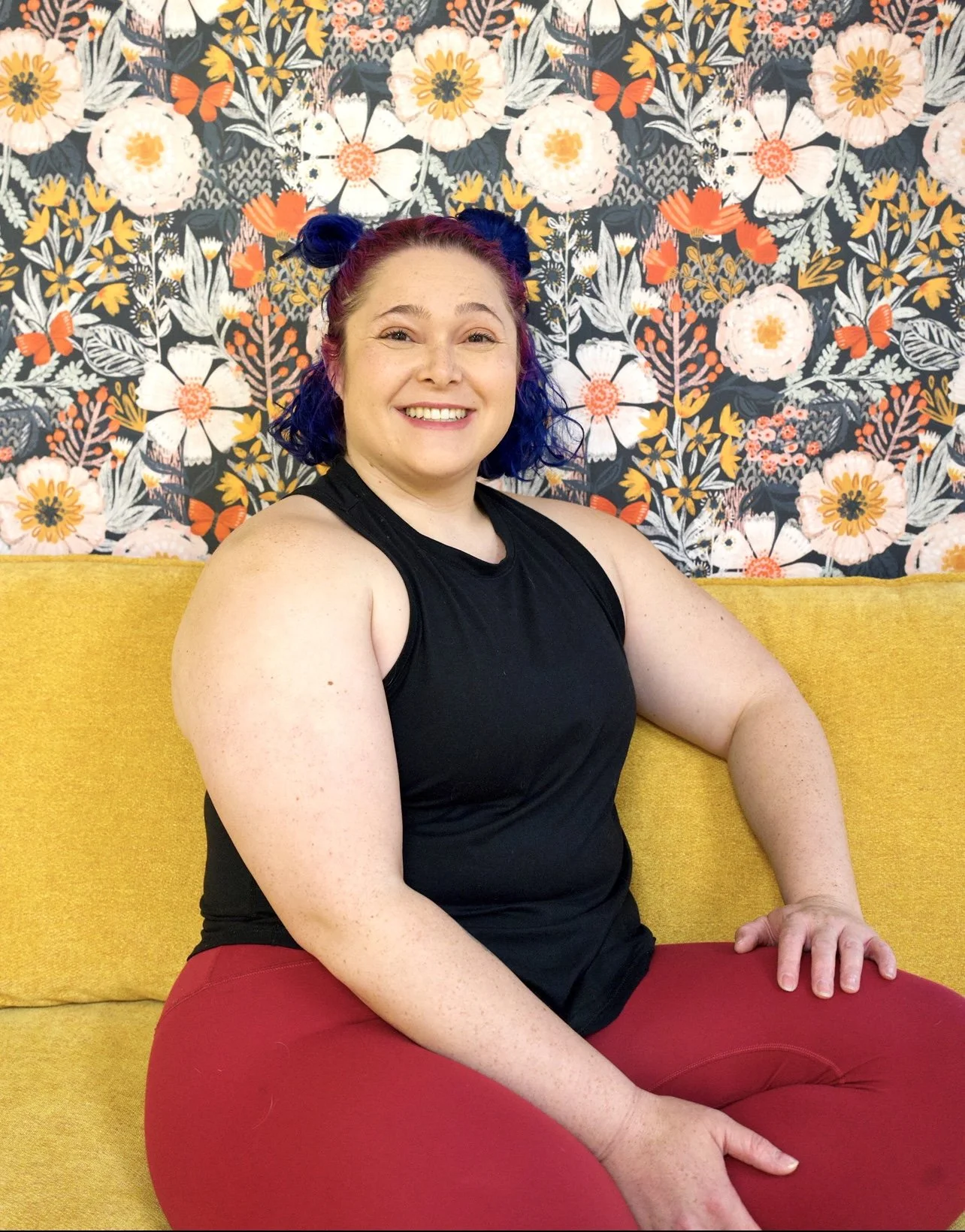 A smiling woman with blue and purple hair styled in two buns, sitting on a yellow couch with a floral and butterfly patterned wall behind her, wearing a black sleeveless top and red pants.