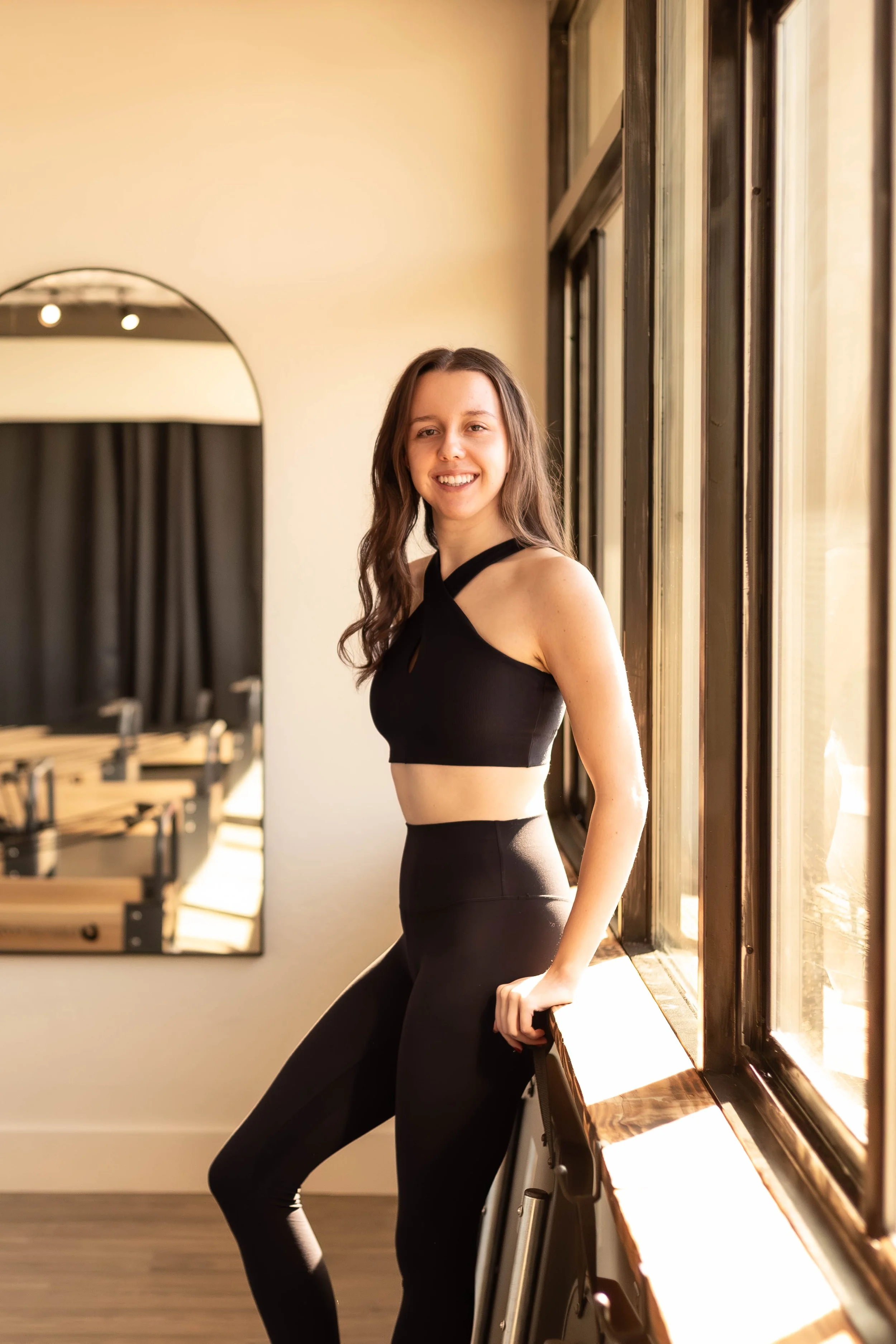 A young woman with brown hair and earrings, wearing a black halter top, smiling, standing against a plain wall.