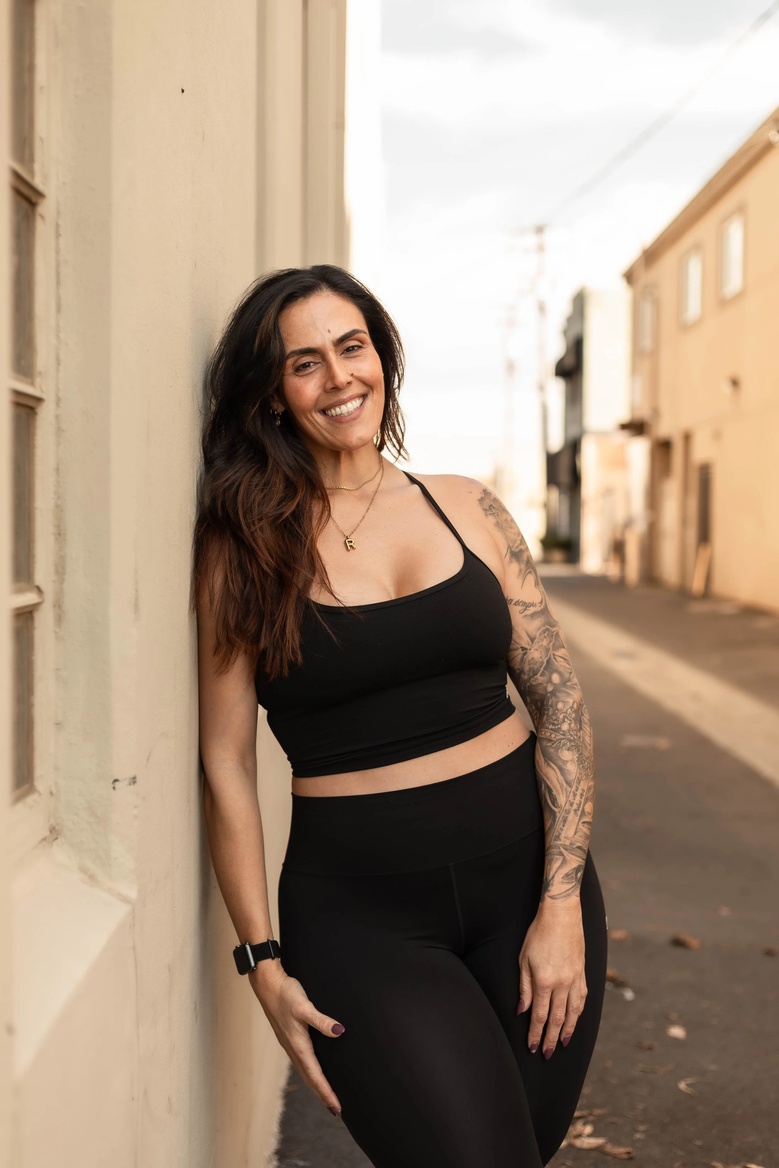 A smiling woman with brown hair tied back, wearing a black sleeveless top, gold jewelry, and displaying a tattoo on her left arm, standing against a plain white wall.