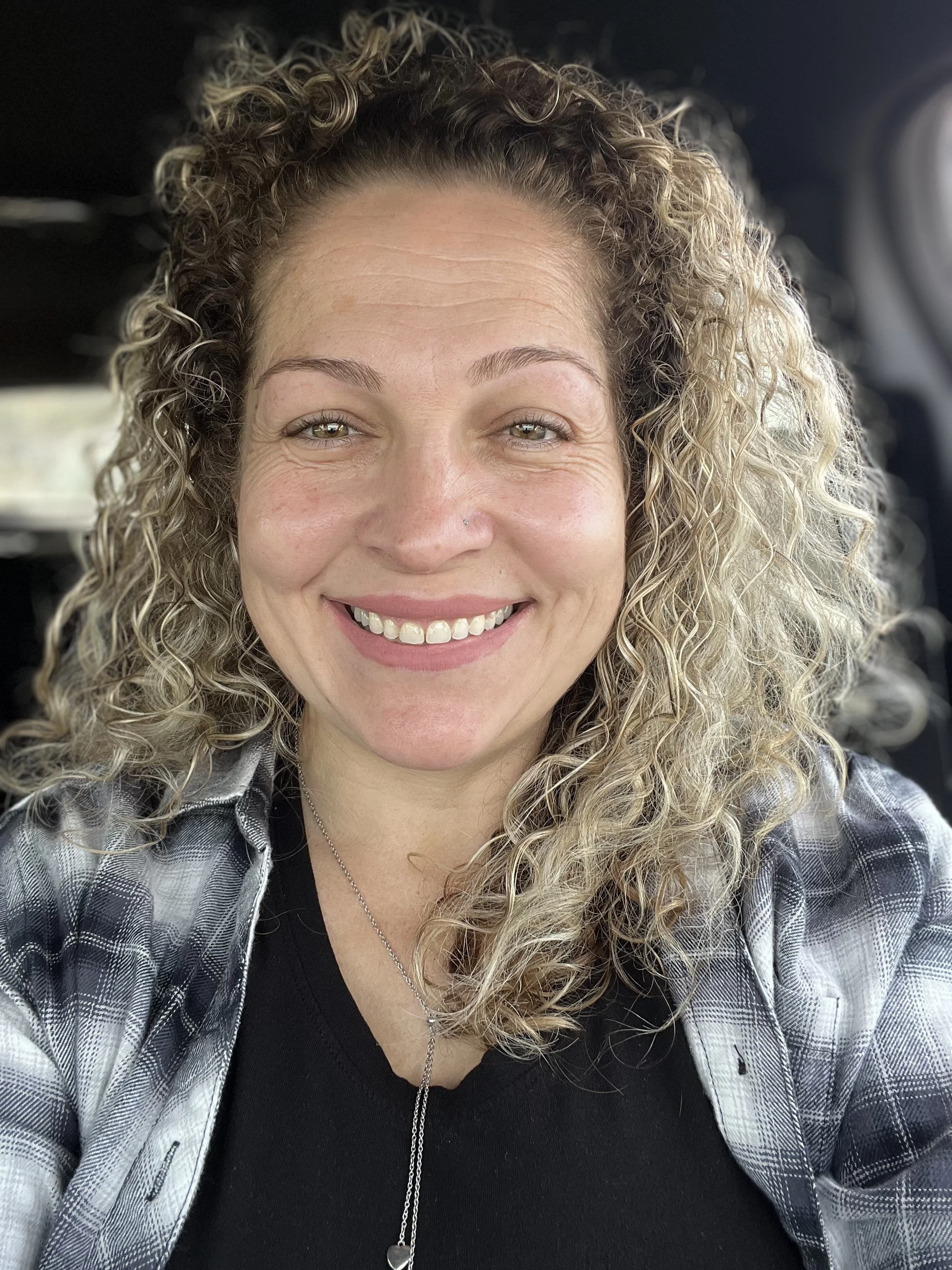 A woman with curly blonde hair smiling at the camera, wearing a black shirt, a checkered shirt, and a necklace, sitting in a vehicle.