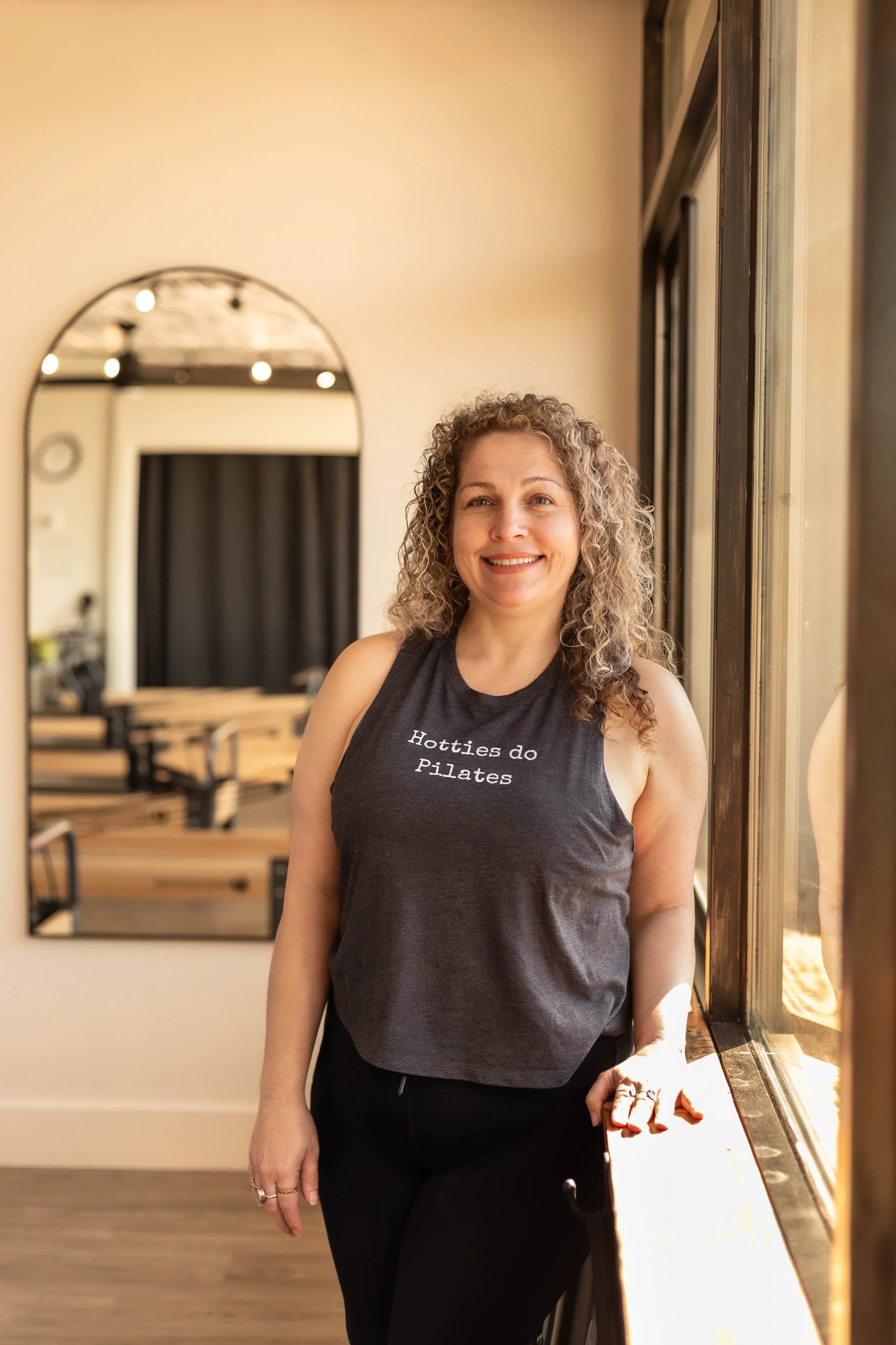 A woman with curly blonde hair smiling at the camera, wearing a black shirt, a checkered shirt, and a necklace, sitting in a vehicle.