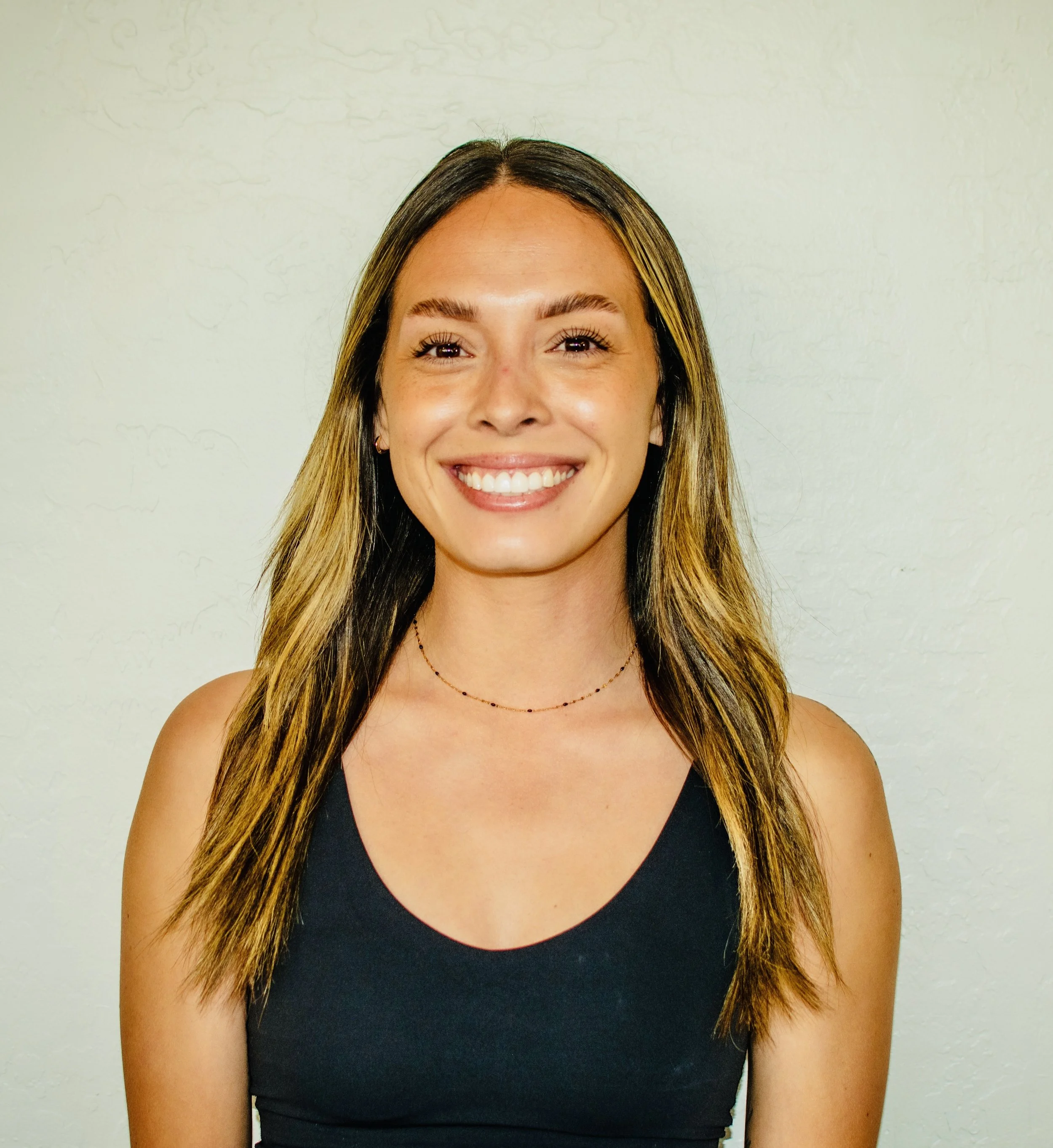 A young woman with long, wavy hair and a bright smile, wearing a black tank top and a delicate necklace, standing against a plain light-colored background.
