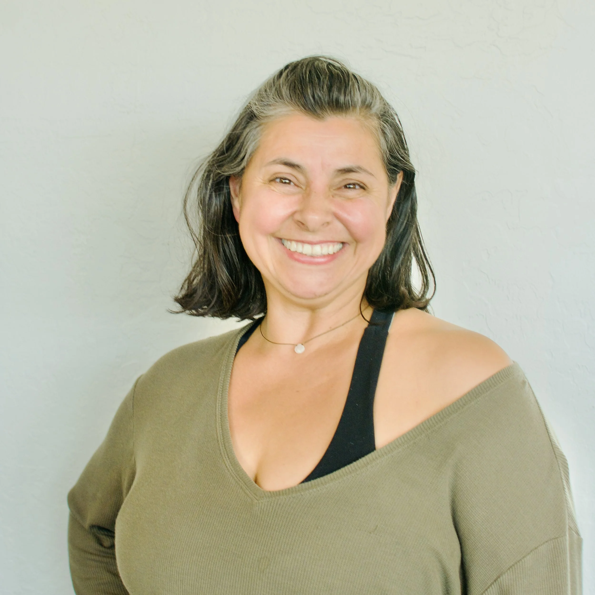 Smiling woman with dark shoulder-length hair, wearing an olive green top and black sports bra, standing against a plain white wall.