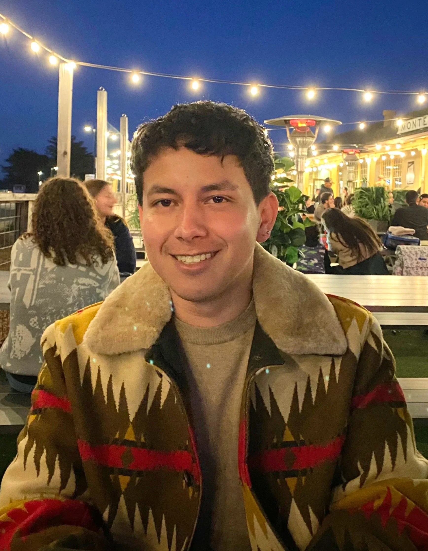 A young man smiling at a restaurant or outdoor dining area during the evening with string lights overhead, surrounded by other people.