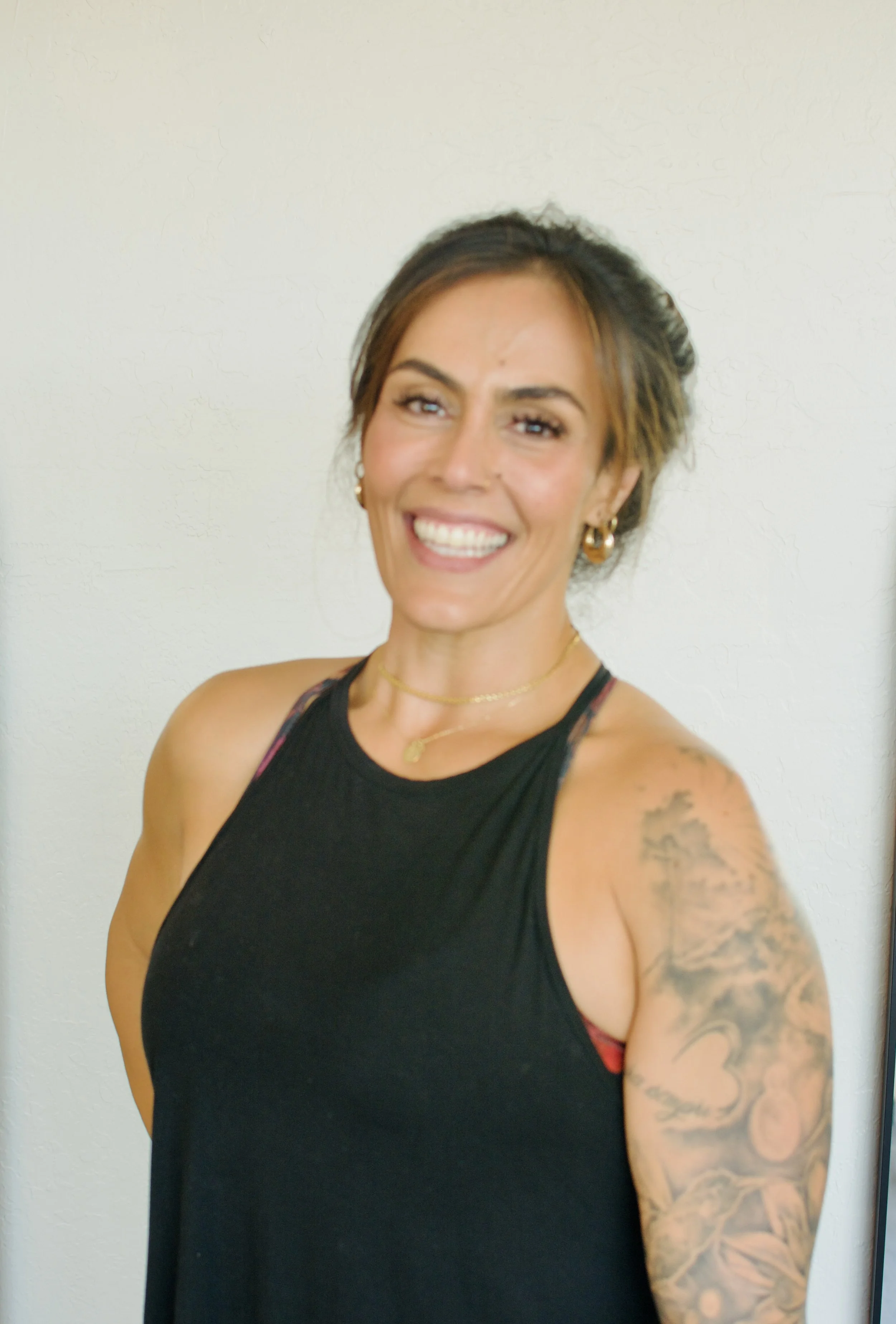 A smiling woman with brown hair tied back, wearing a black sleeveless top, gold jewelry, and displaying a tattoo on her left arm, standing against a plain white wall.