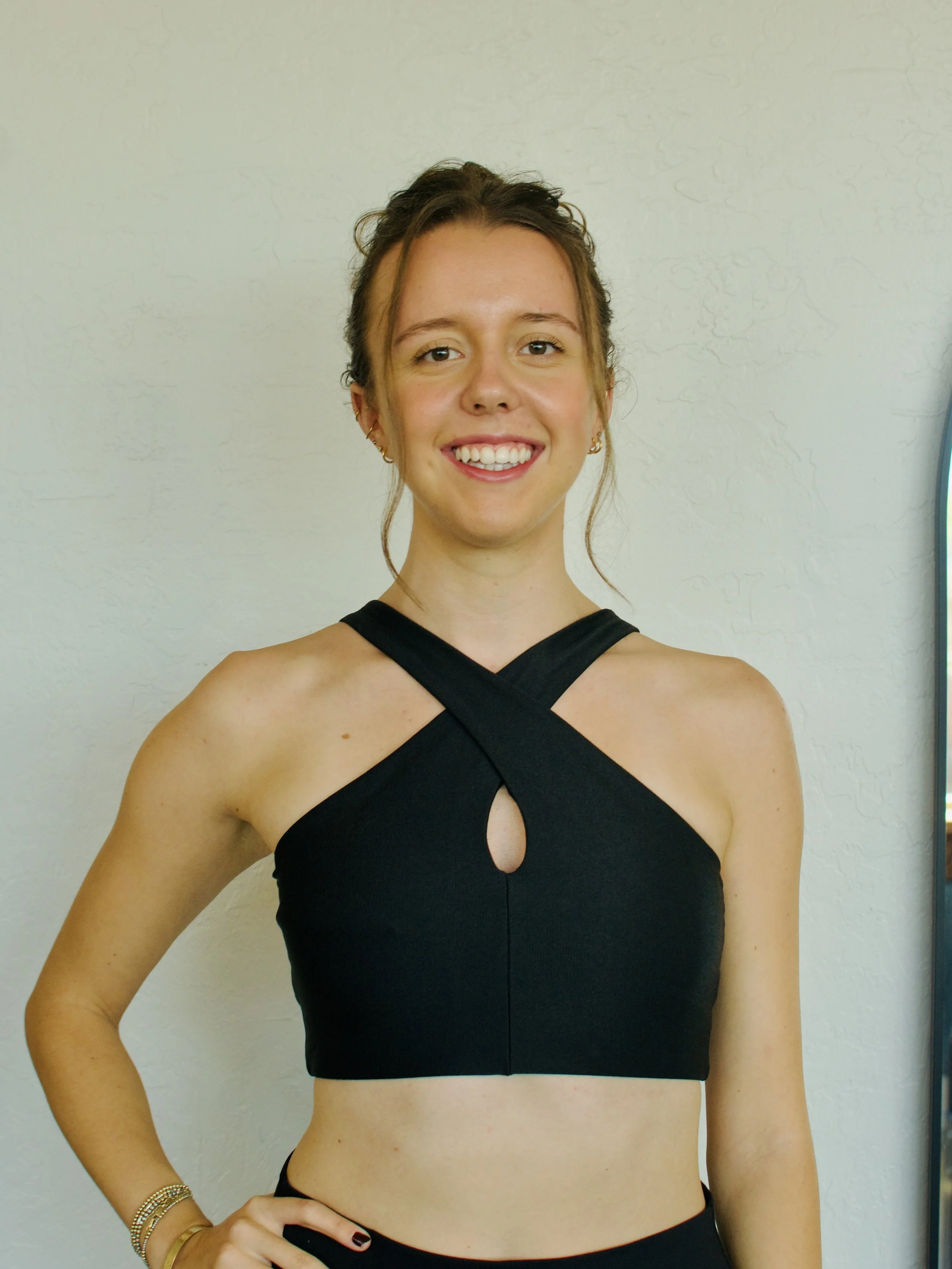 A young woman with brown hair and earrings, wearing a black halter top, smiling, standing against a plain wall.
