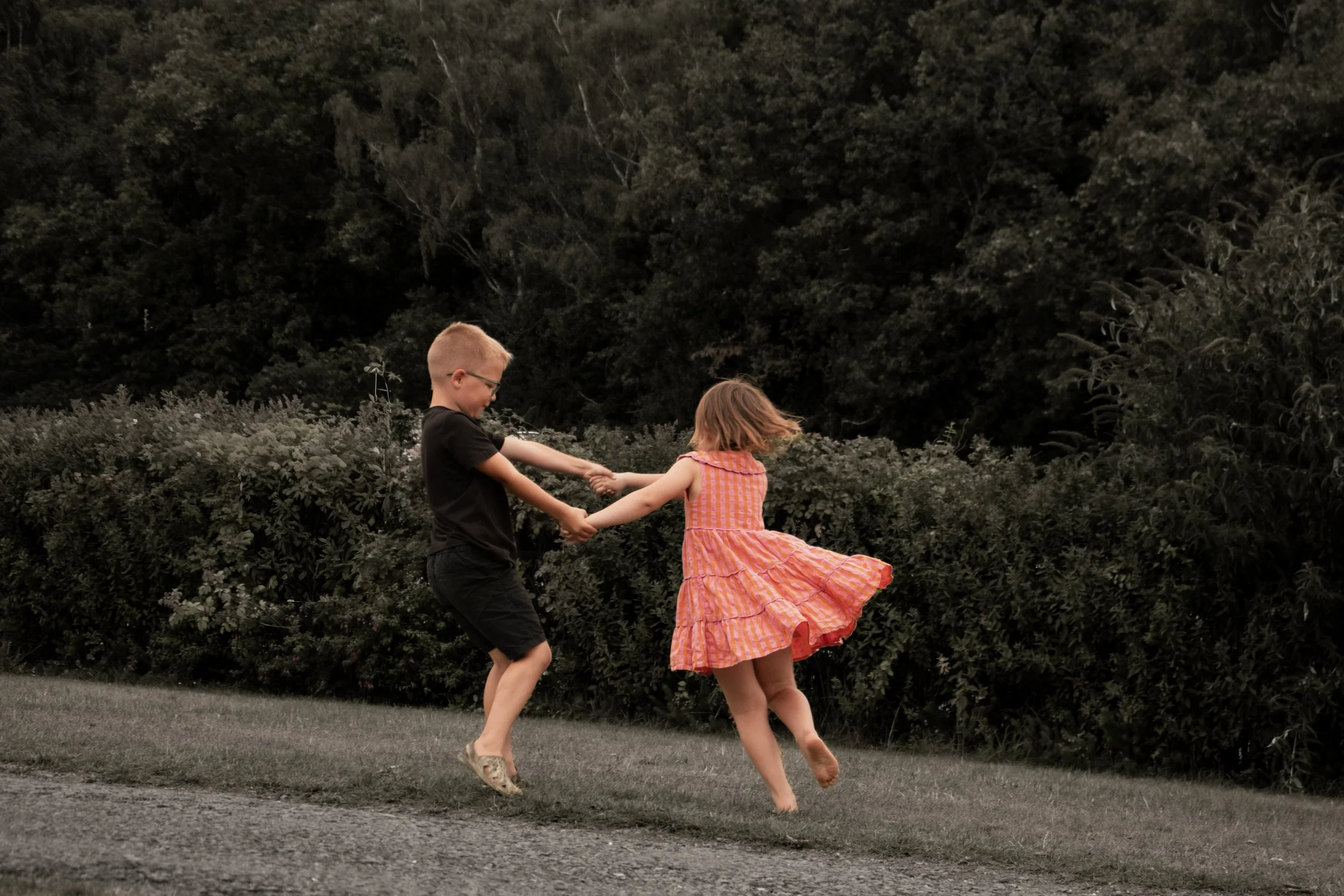 Two children, a boy and a girl, are playing outside holding hands and spinning or dancing on the grass with foliage in the background.