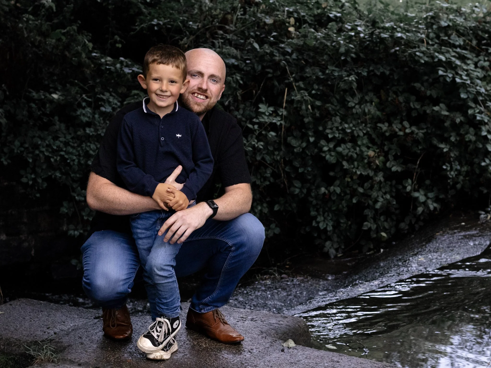 A man and a boy posing together outdoors near a stream, with trees and bushes in the background.