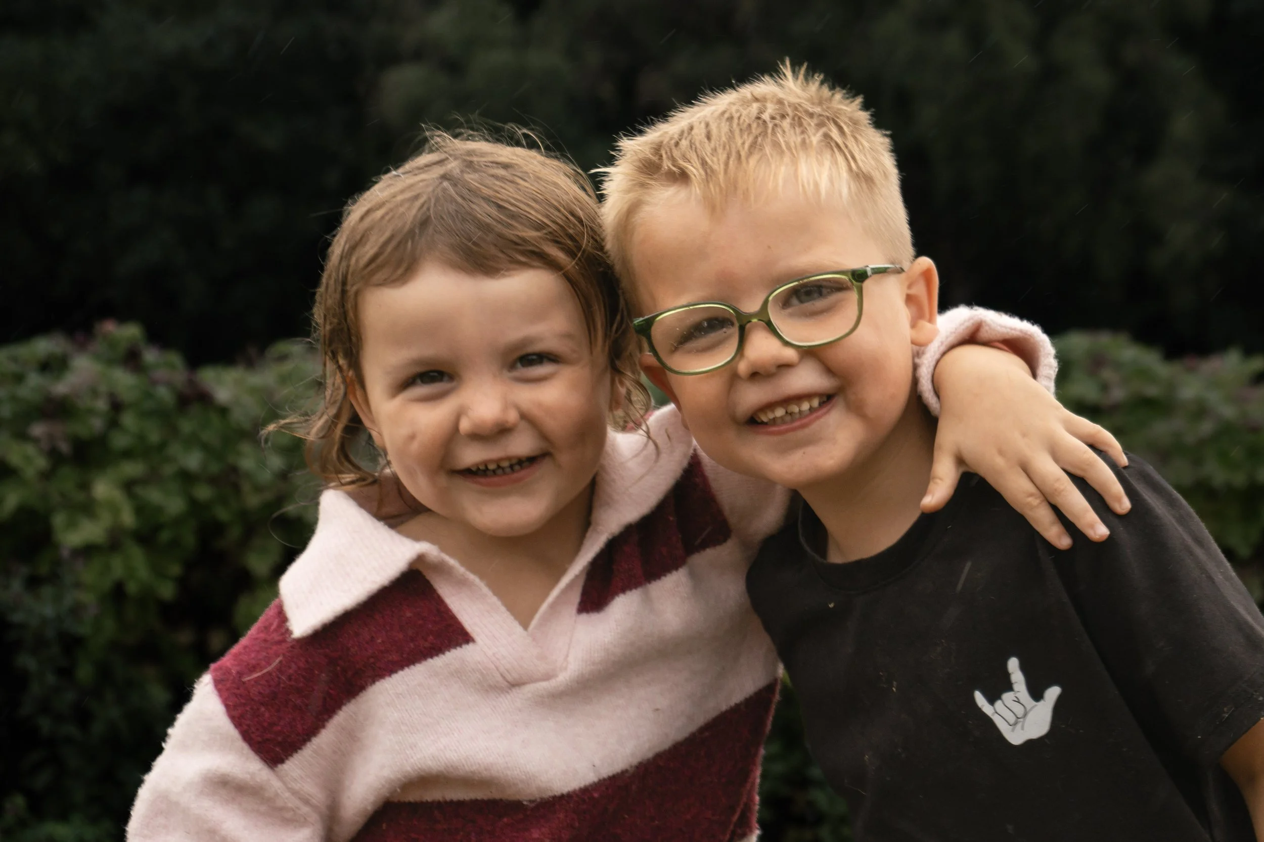 Two young children, a girl and boy, smiling and hugging outdoors with greenery in the background.