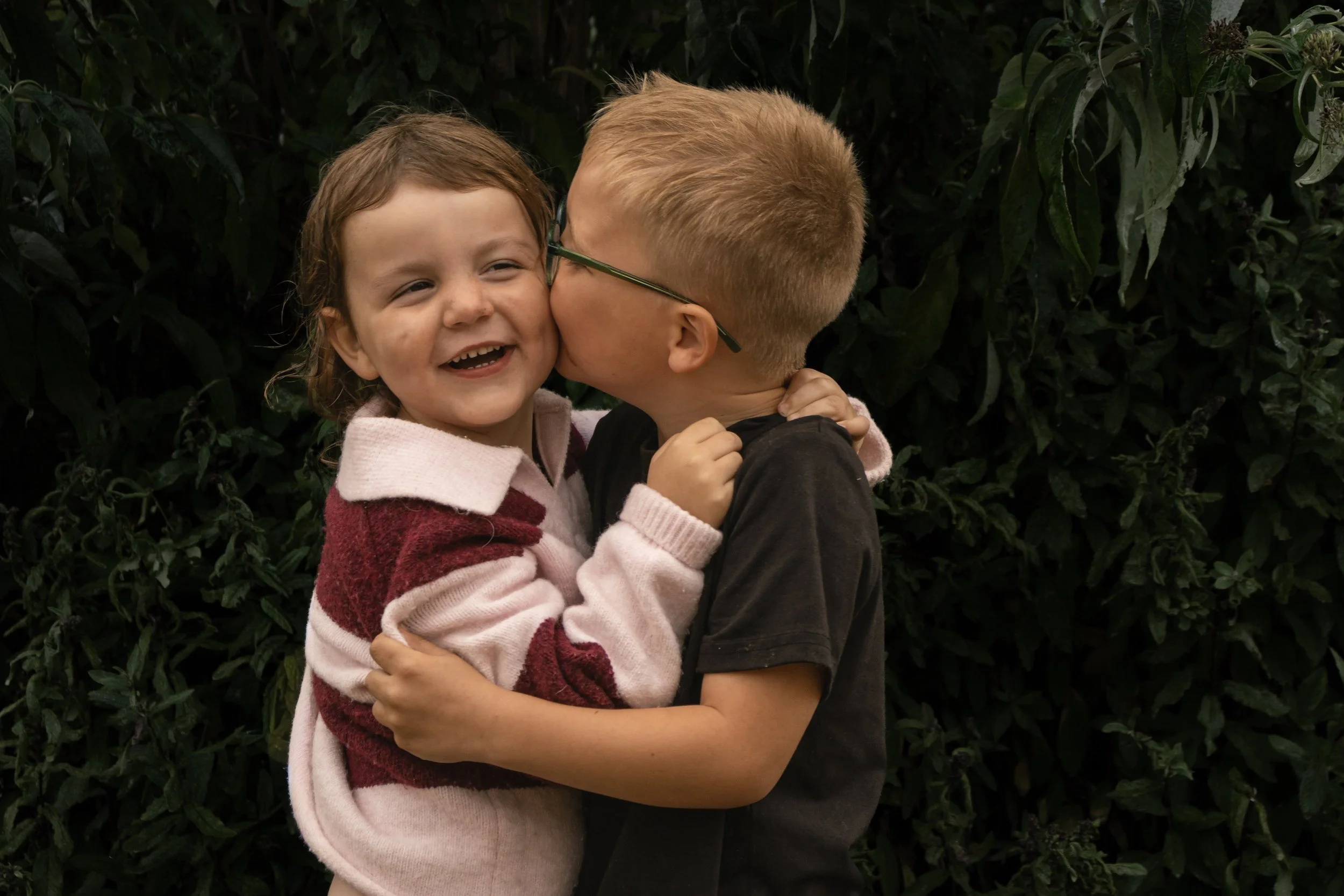 Two children, a girl and a boy, embrace and share a kiss on the cheek, standing in front of a dark leafy background.
