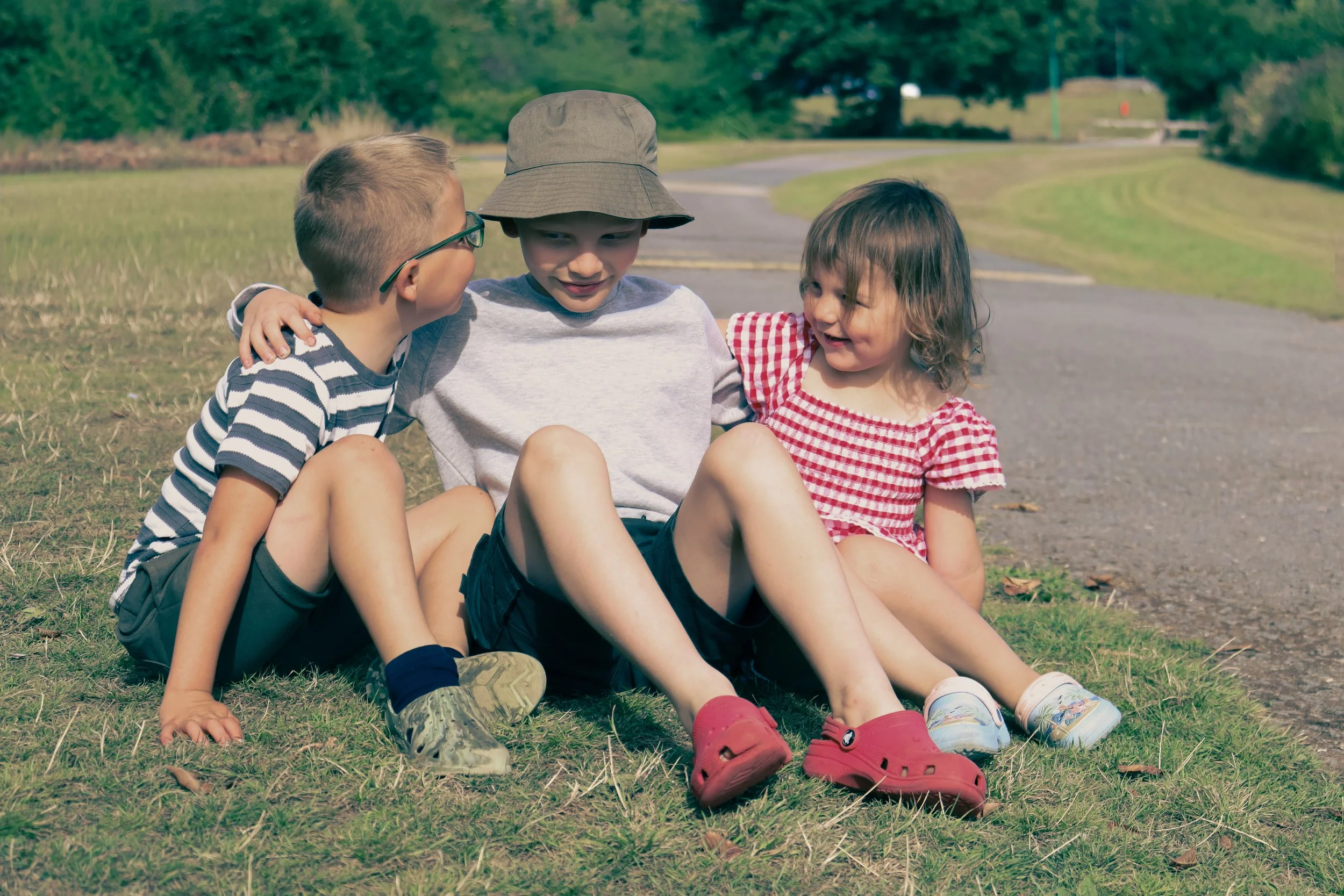 Three children sitting on grass near a path, smiling and interacting with each other on a sunny day.
