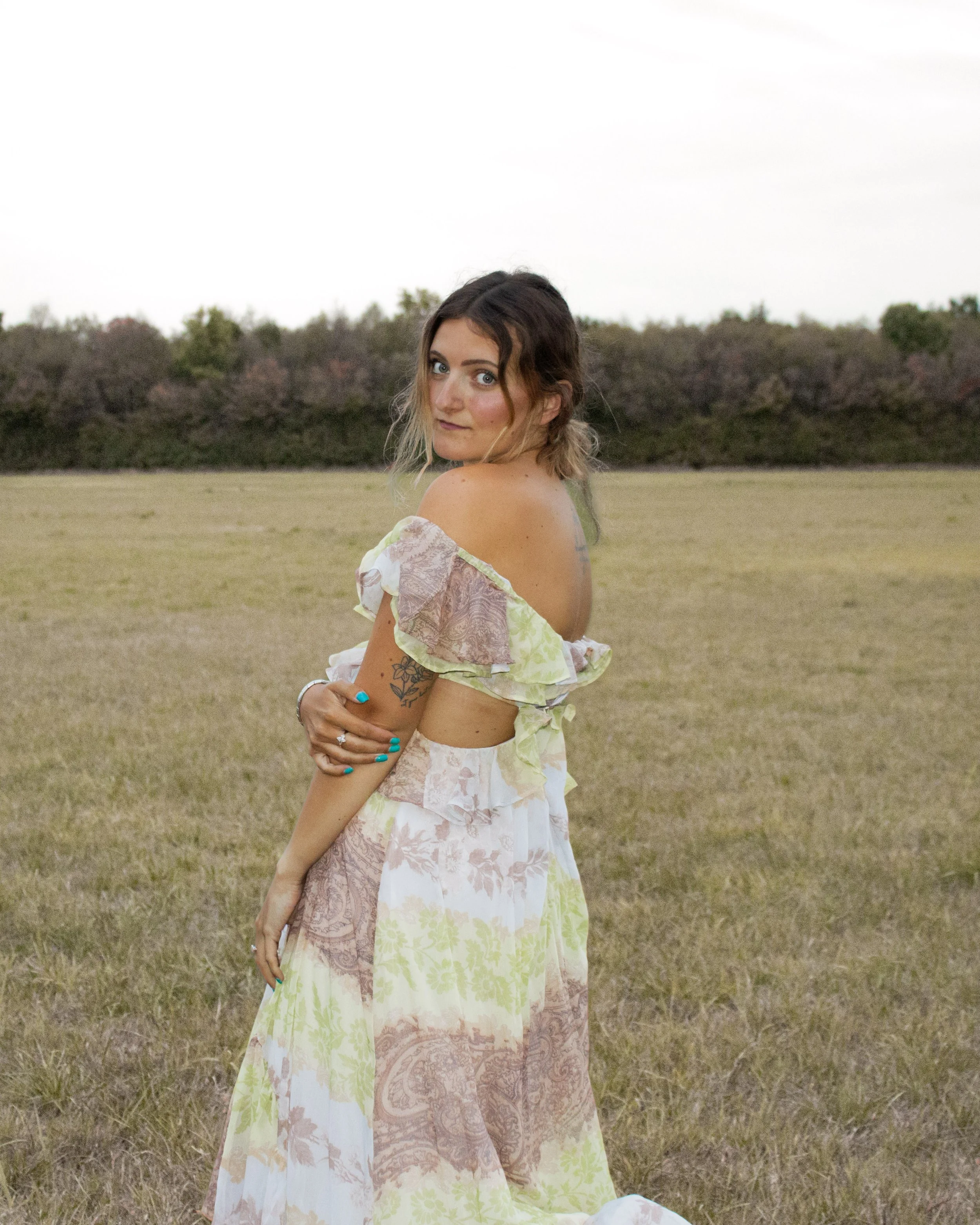 A young woman with wavy hair standing in an open grassy field with trees in the background, wearing a cut-out, off-the-shoulder dress with pastel floral patterns.