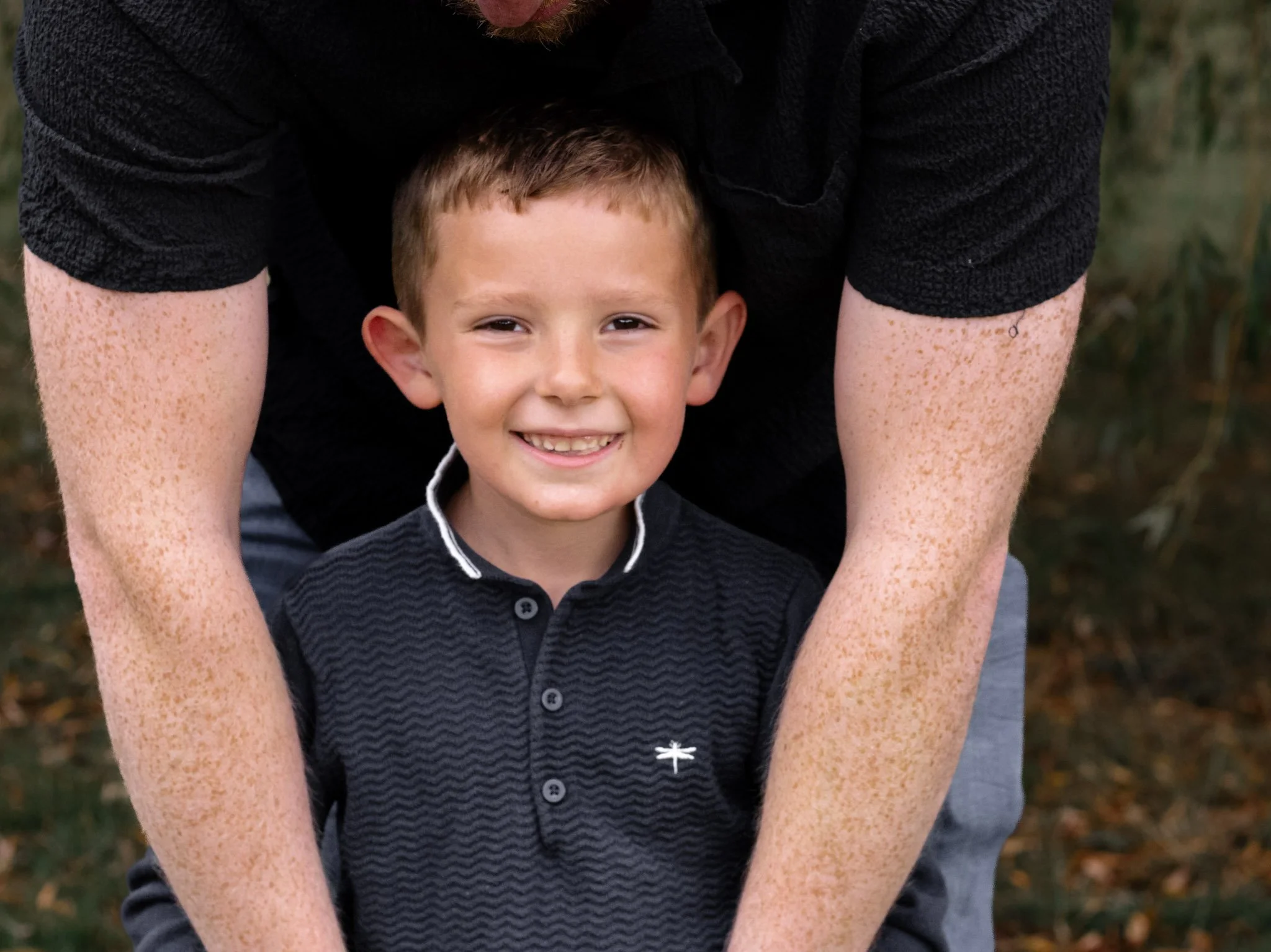 A smiling young boy with short light brown hair, wearing a dark polo shirt with a small logo, standing outdoors with some greenery and fallen leaves in the background.