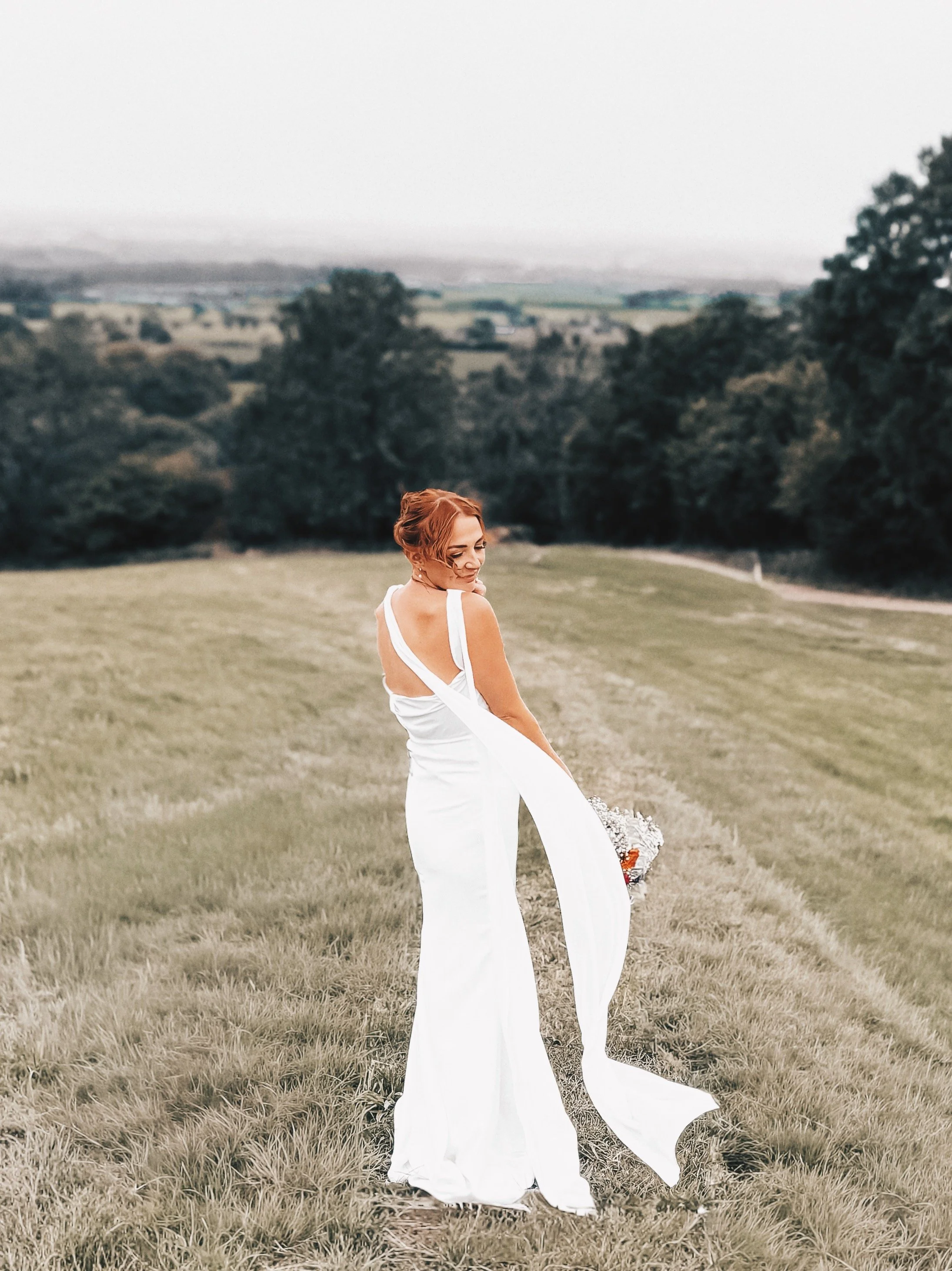 A woman in a white wedding dress stands on a grassy field holding a bouquet, with a backdrop of trees and distant hills.