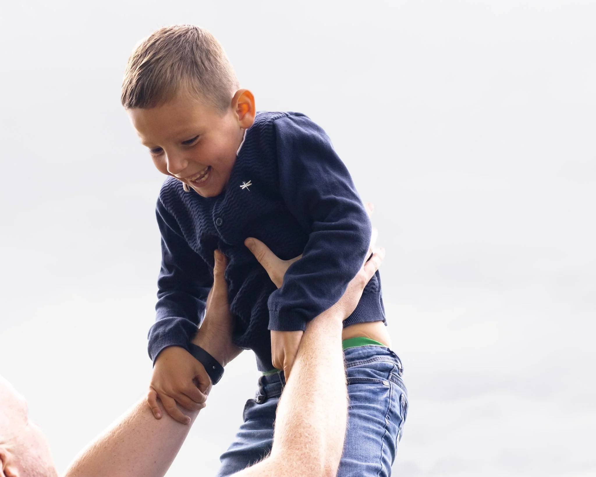 A young boy in a navy sweater and jeans being lifted by an adult, smiling and looking down, outdoors under a cloudy sky.