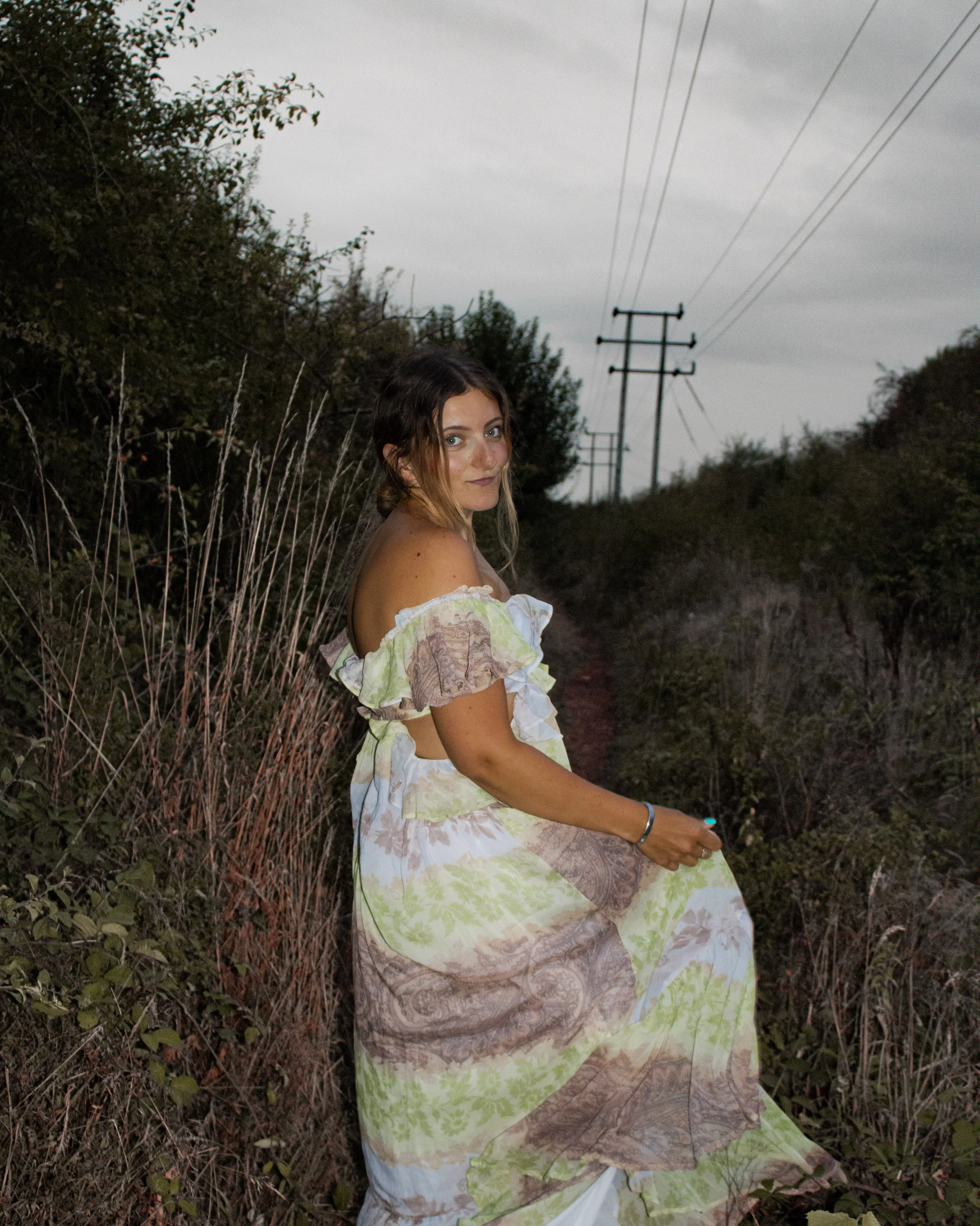 A woman in a colorful off-shoulder dress sitting on a grassy hillside with power lines in the background during dusk.