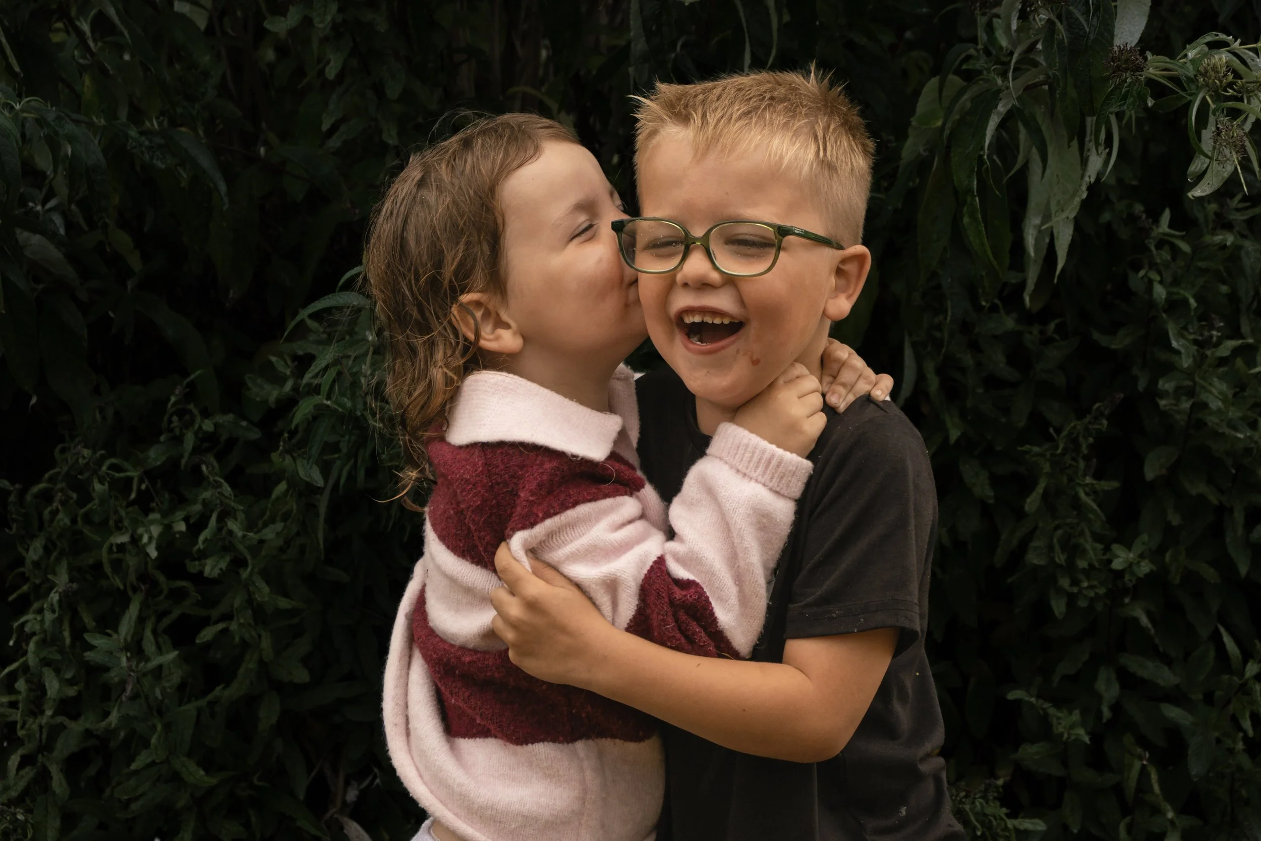 A young girl is kissing a young boy on the cheek while they embrace, standing in front of dark green foliage. The boy is wearing glasses, a black shirt, and has a joyful expression.