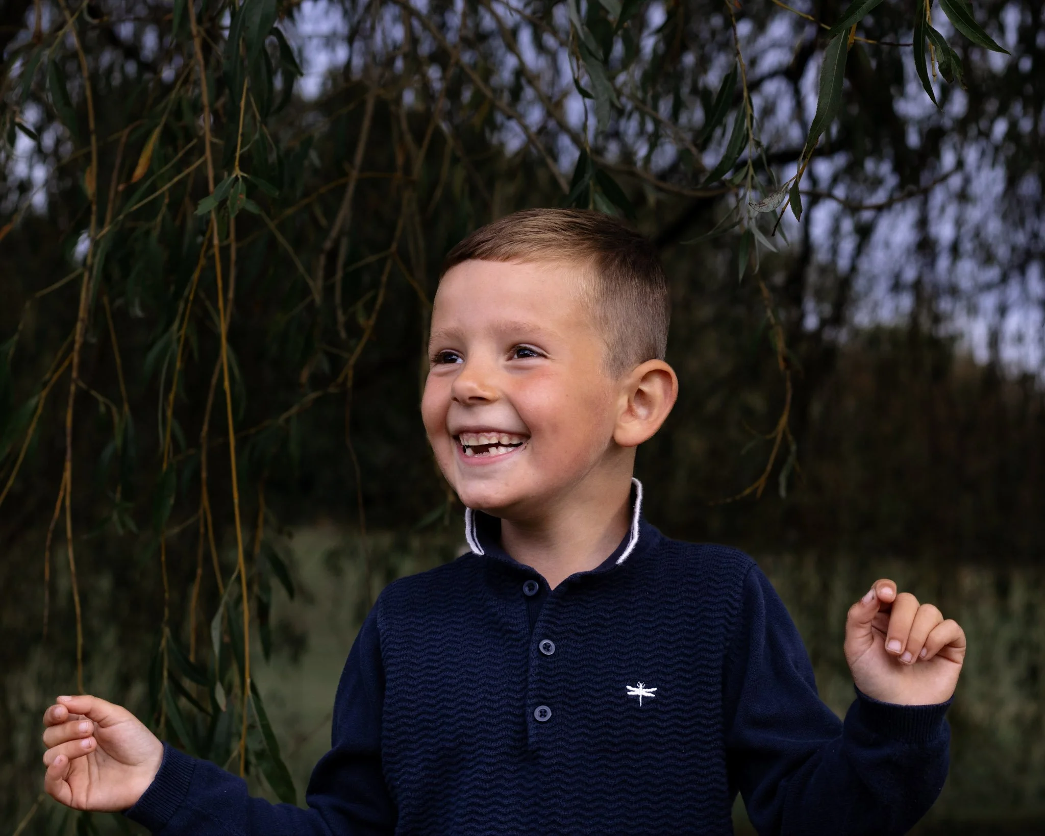 A young boy with short light brown hair, smiling happily, wearing a navy blue shirt, standing outdoors near a tree with long, hanging leaves.