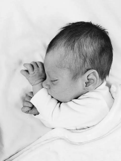 A sleeping baby with short dark hair, curled up on a soft surface, wearing a long sleeve shirt.