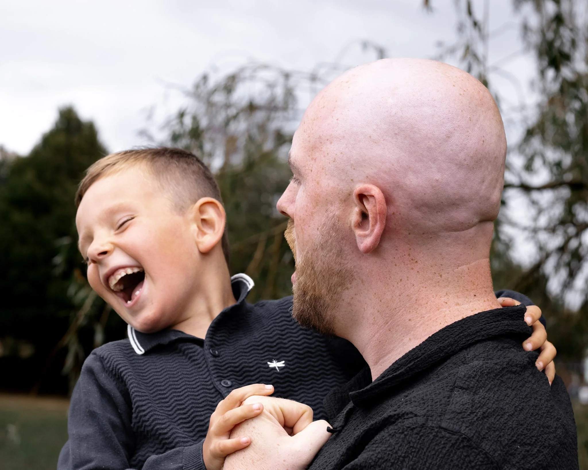 A child and an adult enjoying a joyful moment outdoors, with the child laughing and the adult holding him, against a background of trees and cloudy sky.