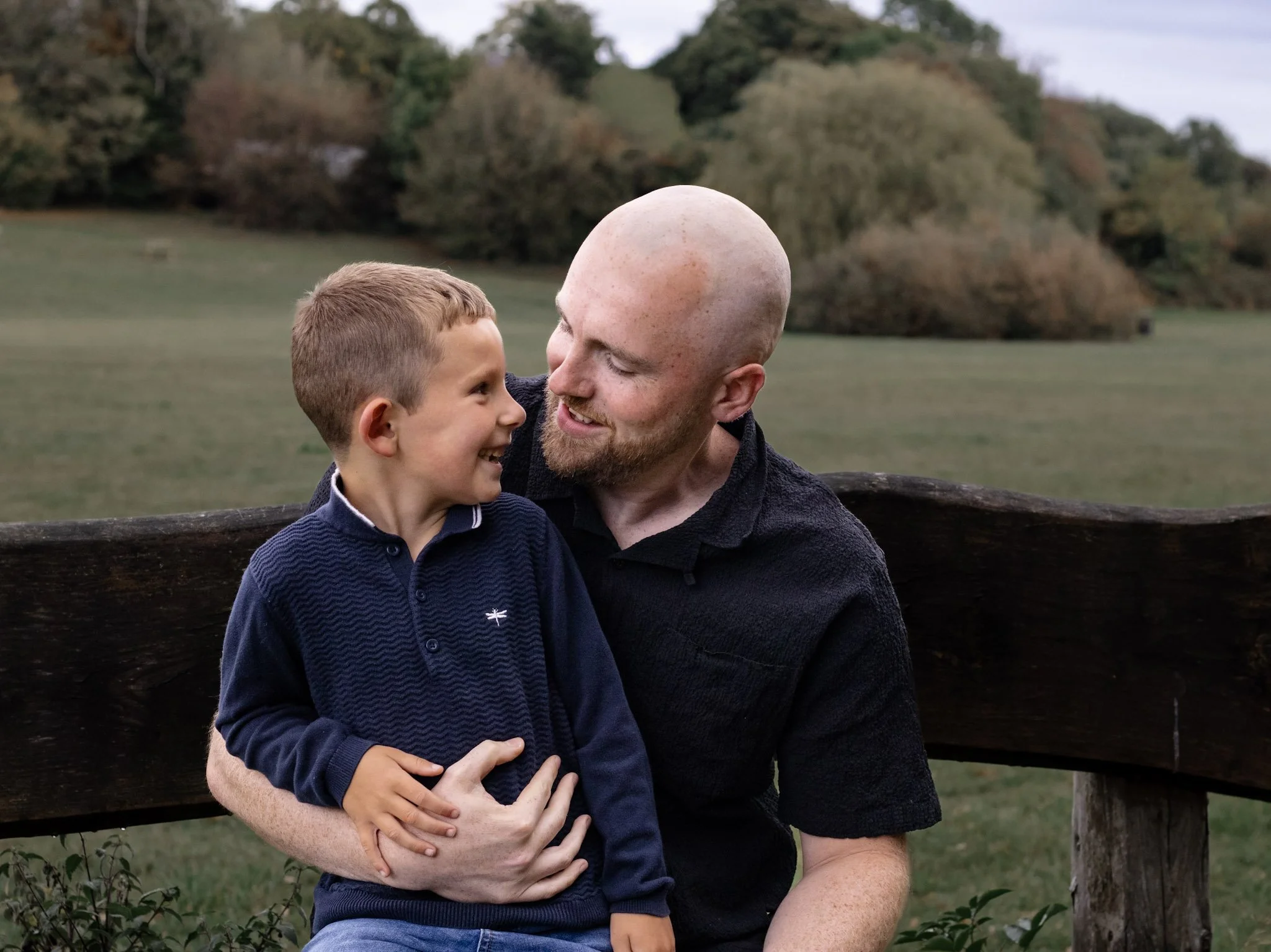 A man and a young boy smiling at each other outdoors near a wooden fence with trees in the background.
