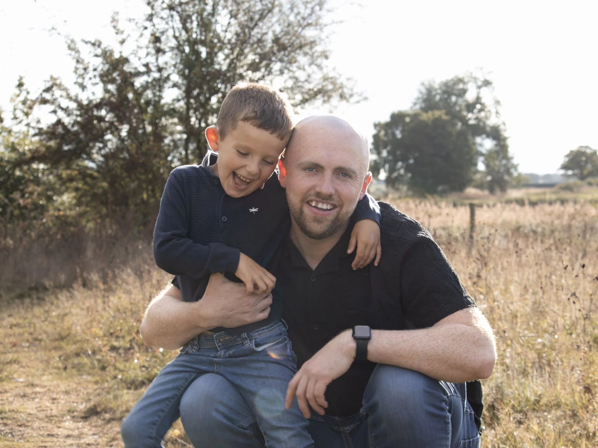 A man and boy smiling outdoors in a field with trees in the background, enjoying a sunny day.