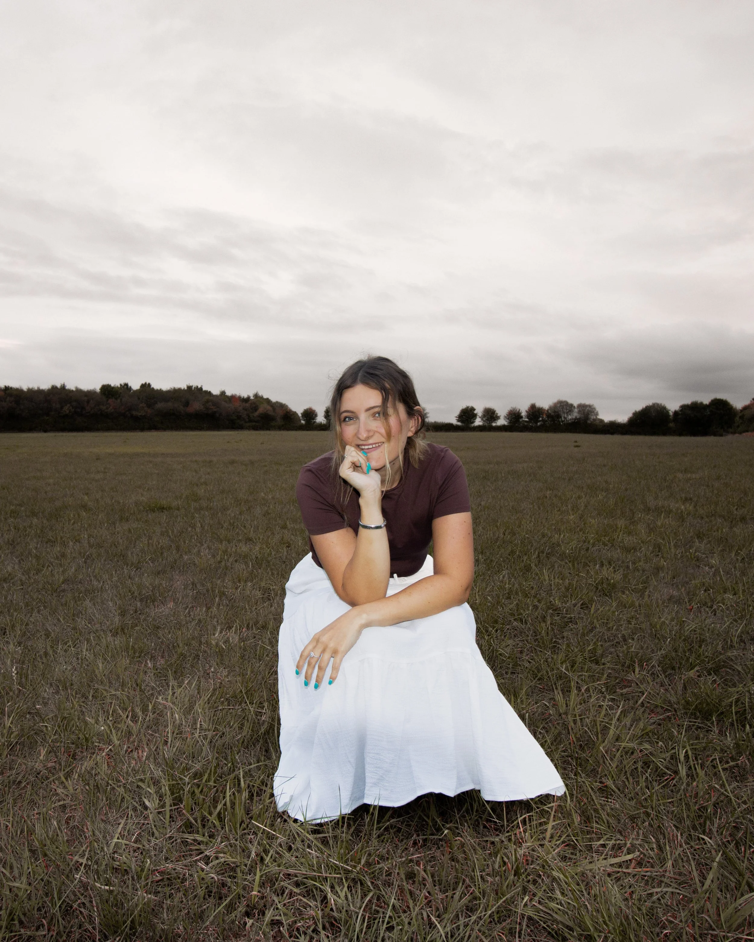 Woman sitting on grassy field, smiling, with overcast sky and trees in background.