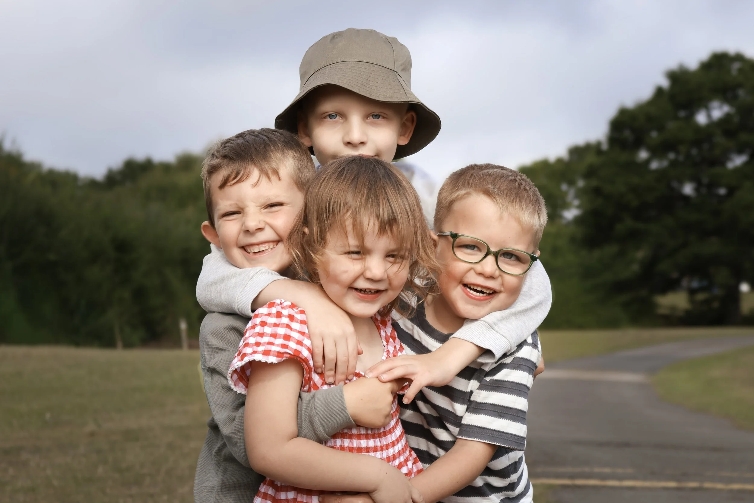 Four children smiling and hugging outdoors on a cloudy day with trees and a pathway in the background.