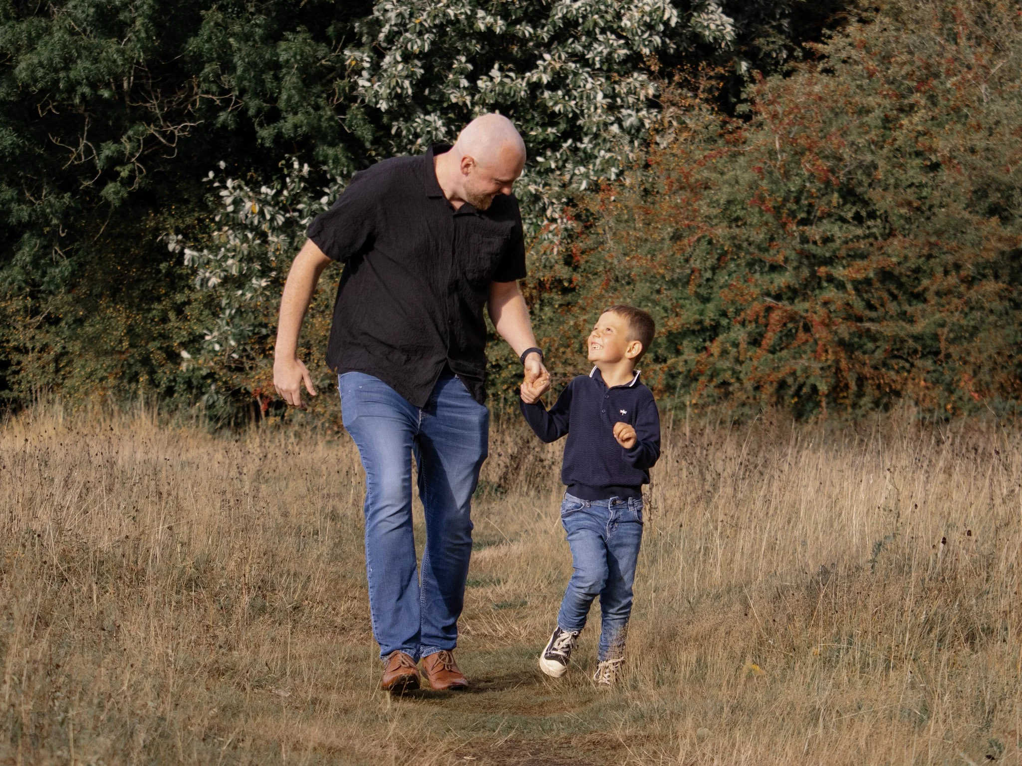 A man and a young boy walk hand in hand through a grassy field with trees in the background. The man is bald, wearing a black shirt and blue jeans. The boy is smiling, wearing a navy sweater and jeans.