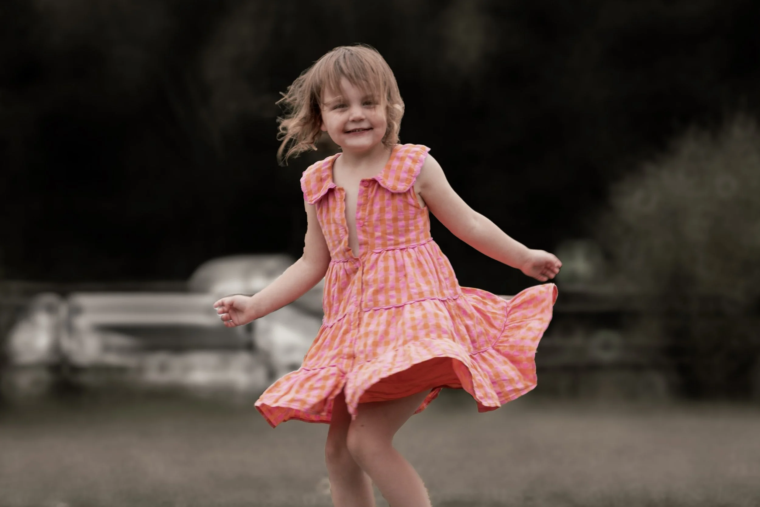 A young girl in a pink and orange checkered dress twirling outdoors on a paved path with blurred trees and vehicles in the background.