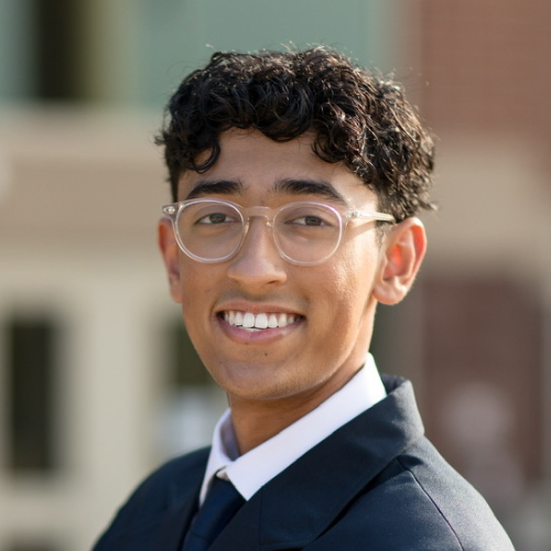 Young man with curly hair and glasses smiling outdoors in professional attire.
