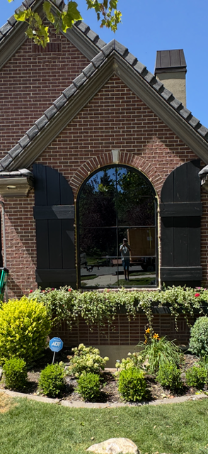 Reflection of a person in a large arched window on a brick house with black shutters, surrounded by a garden with bushes and flowers.