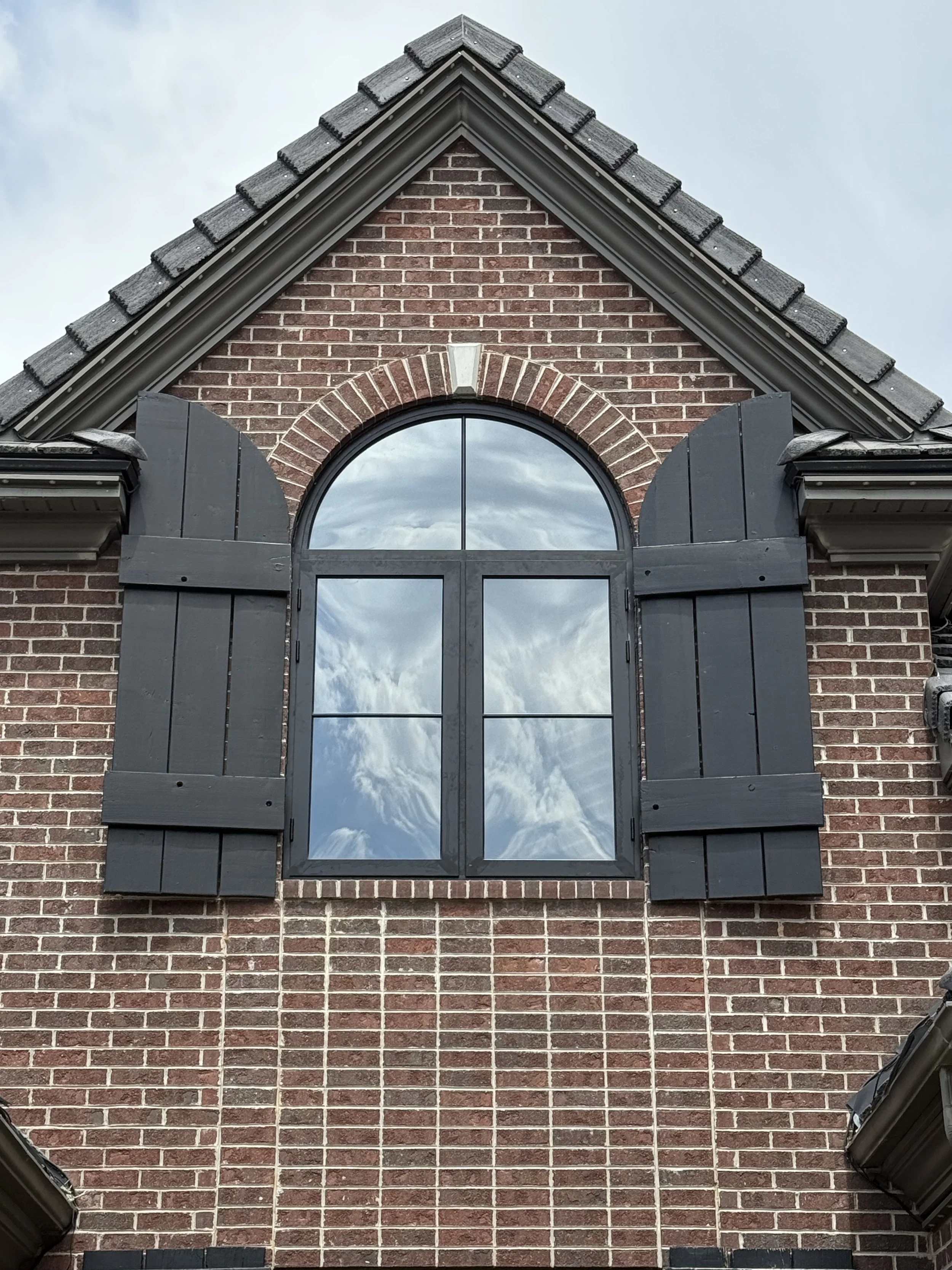 Brick house with large arched window and black shutters, reflecting cloudy sky