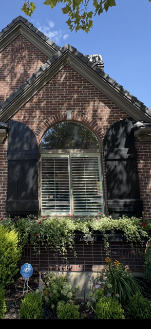 Front view of a brick house with an arched window, black shutters, and a landscape garden with various plants and flowers in front.